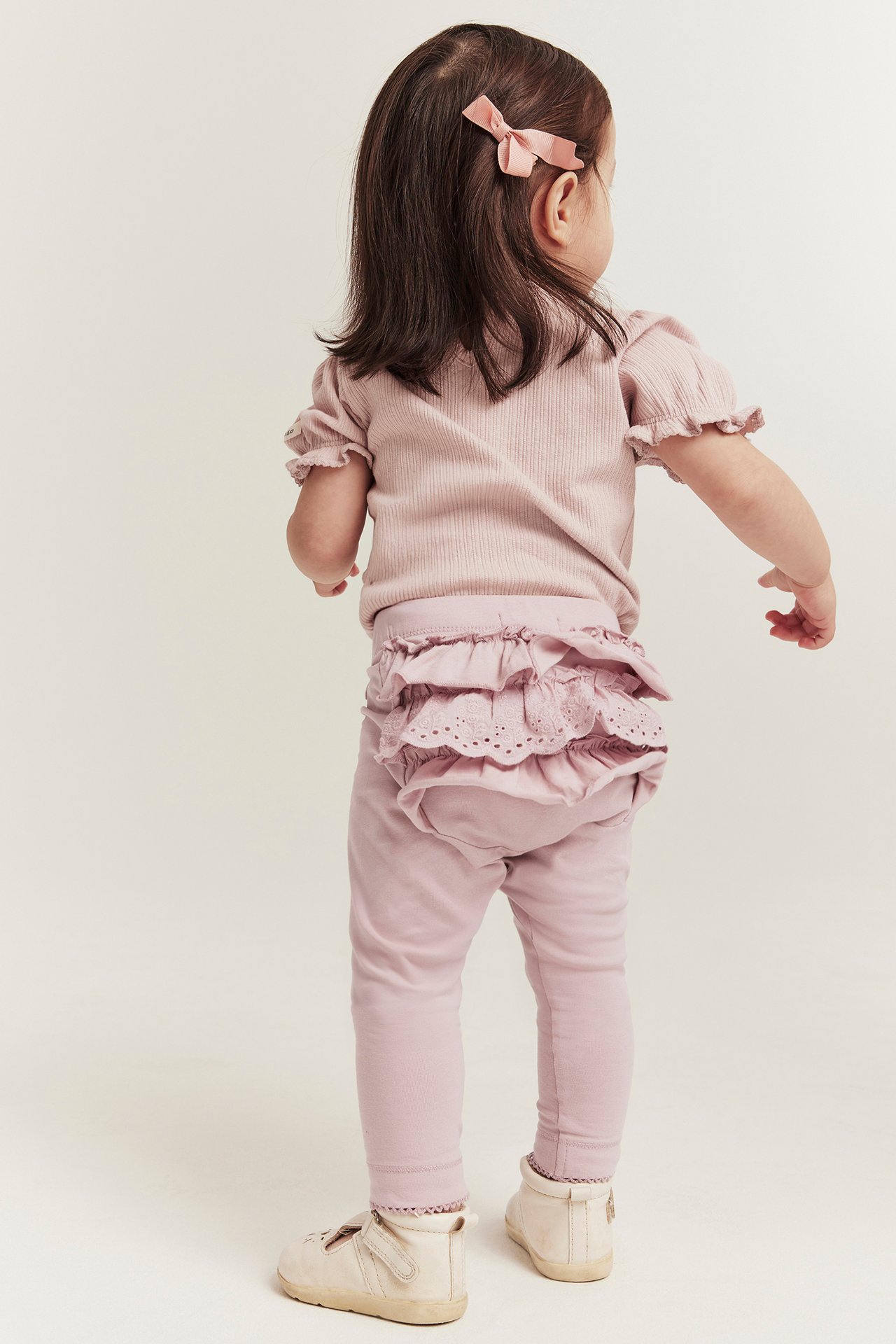 Back view of a baby in pink ruffled leggings with lace, a pink ribbed top, light shoes, and a pink bow.