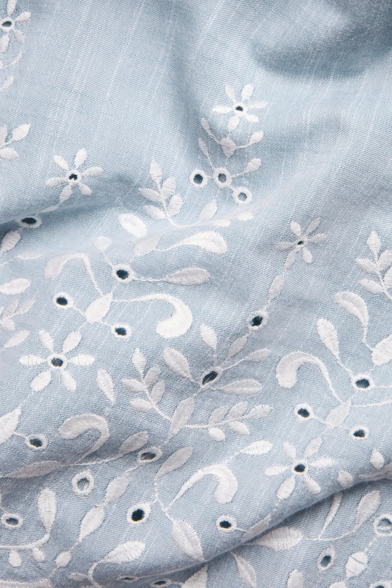 Close-up of light blue cotton chambray fabric with white floral embroidery and cut-out details.