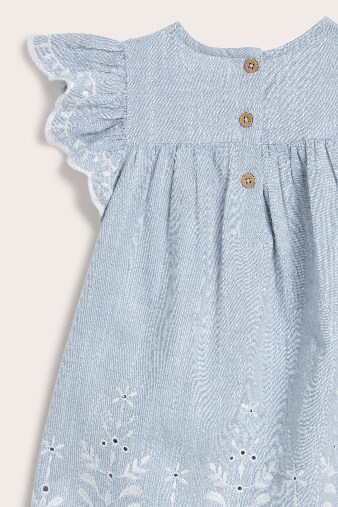 Close-up back view of a light blue chambray kid's dress with white embroidery, ruffled sleeves, and back buttons.