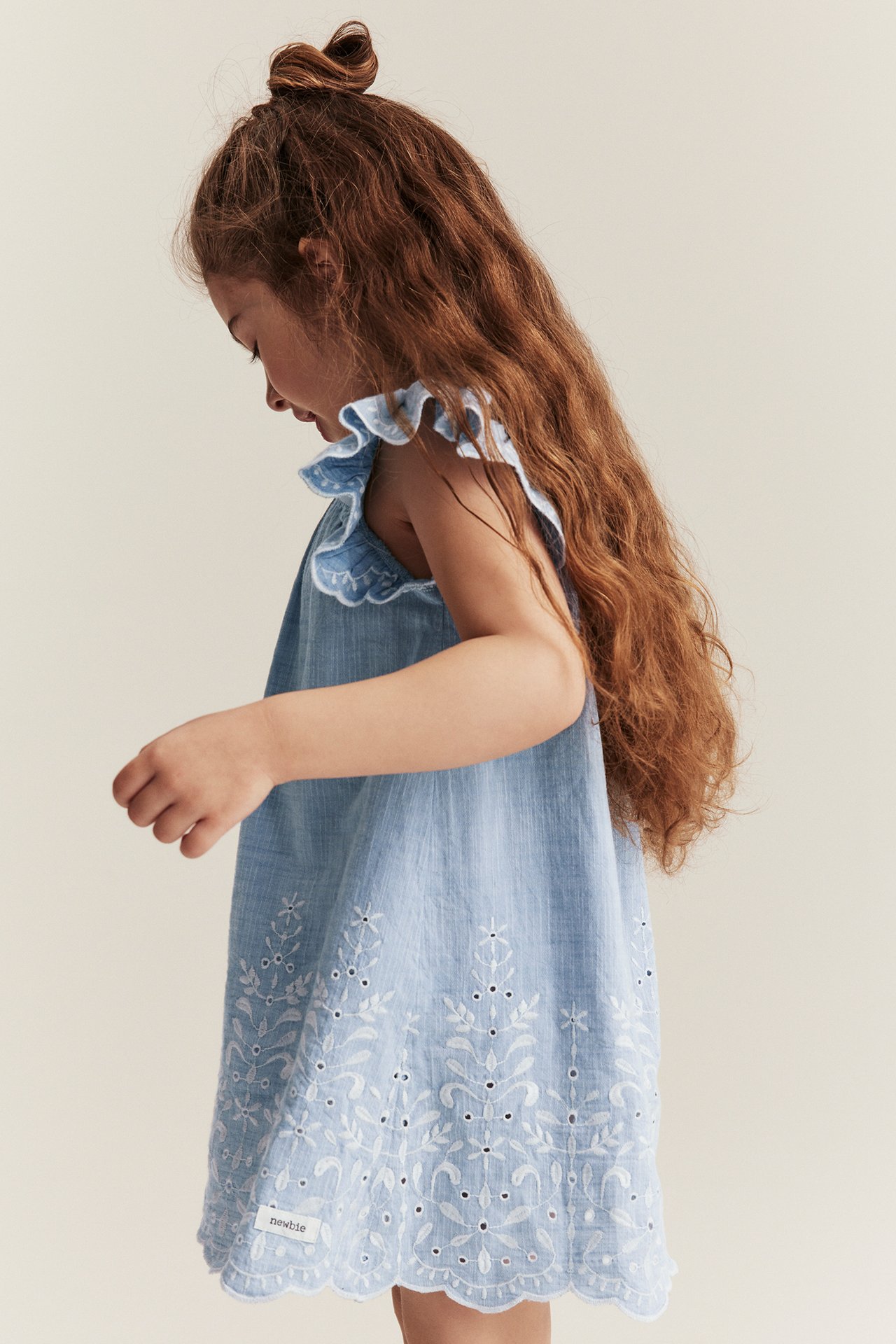 Side view of a kid wearing a light blue chambray dress with white embroidery, ruffled shoulders, and a scalloped hem.