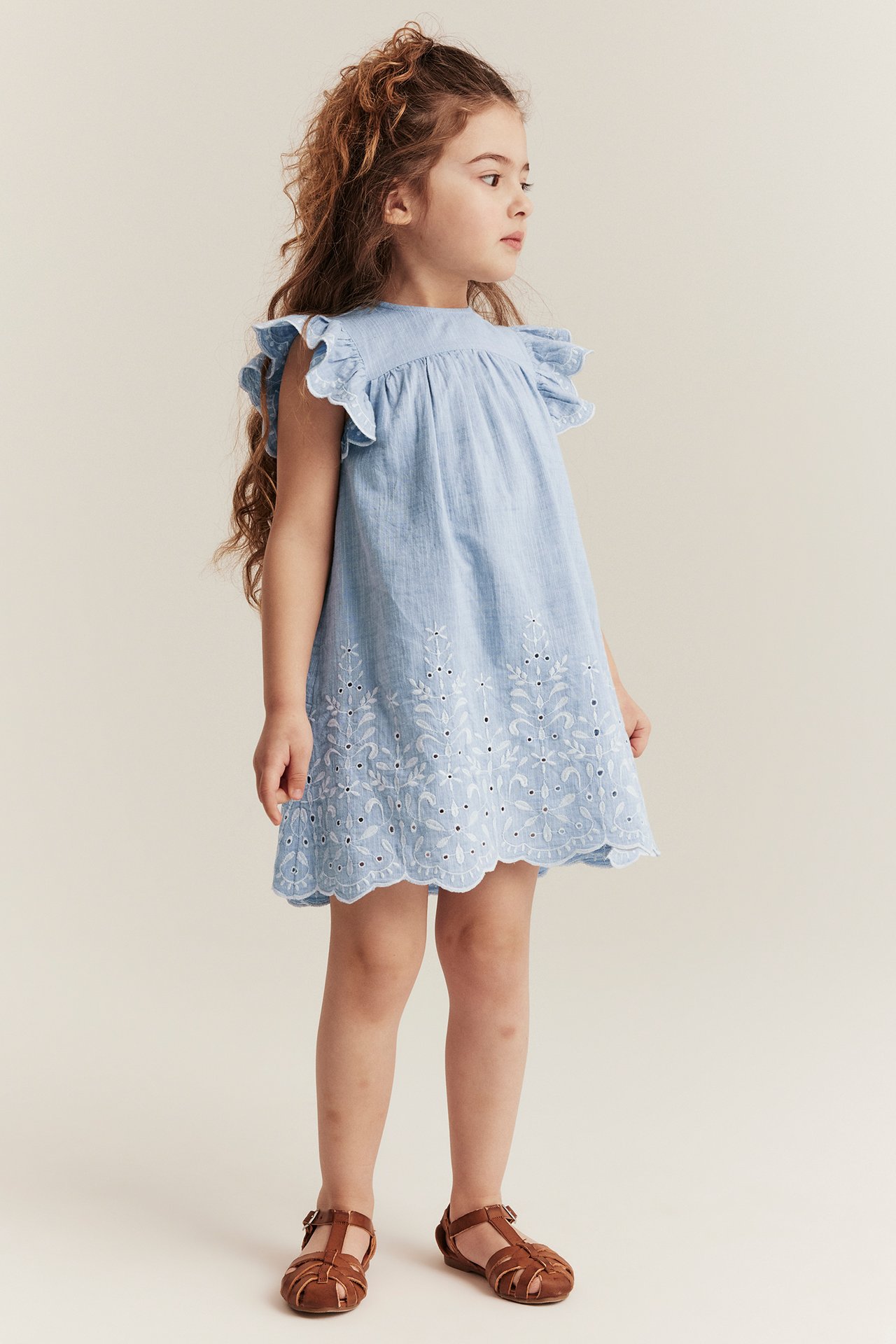 Side view of a kid in a light blue chambray dress with white embroidery and brown sandals.