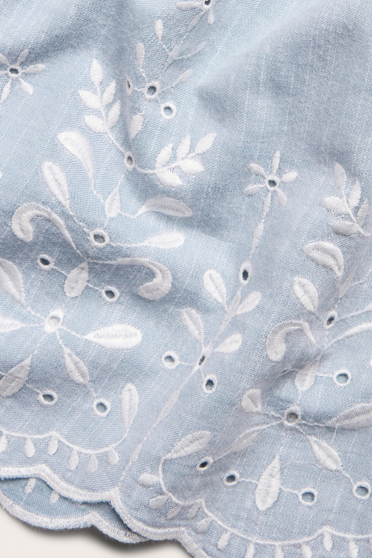 Close-up of a light blue chambray dress with white floral embroidery and a scalloped edge.