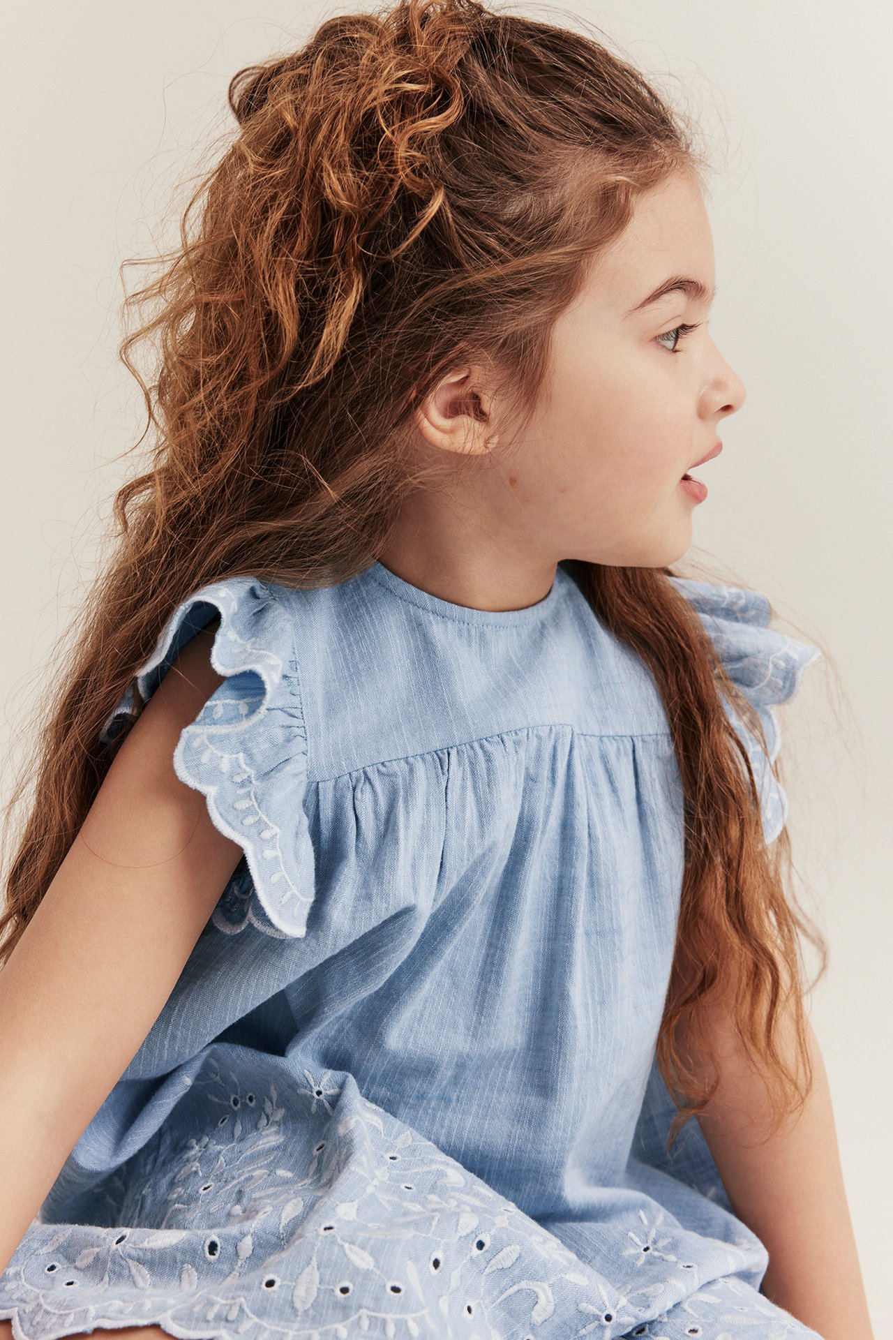 Side view of a kid wearing a light blue chambray dress with white embroidered ruffles and hem.