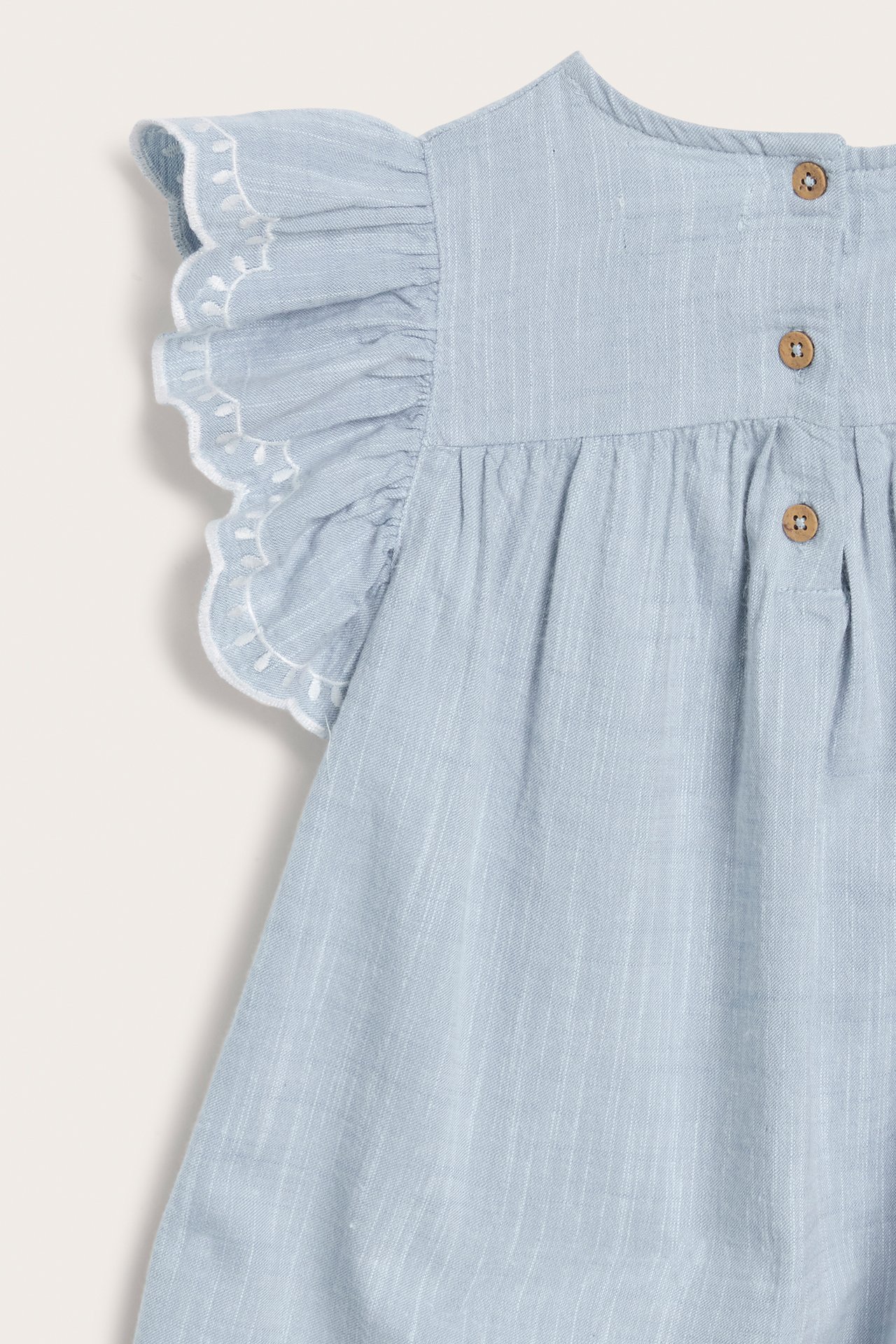 Close-up back view of a light blue chambray kid's dress with white embroidered ruffled sleeves and wooden buttons.