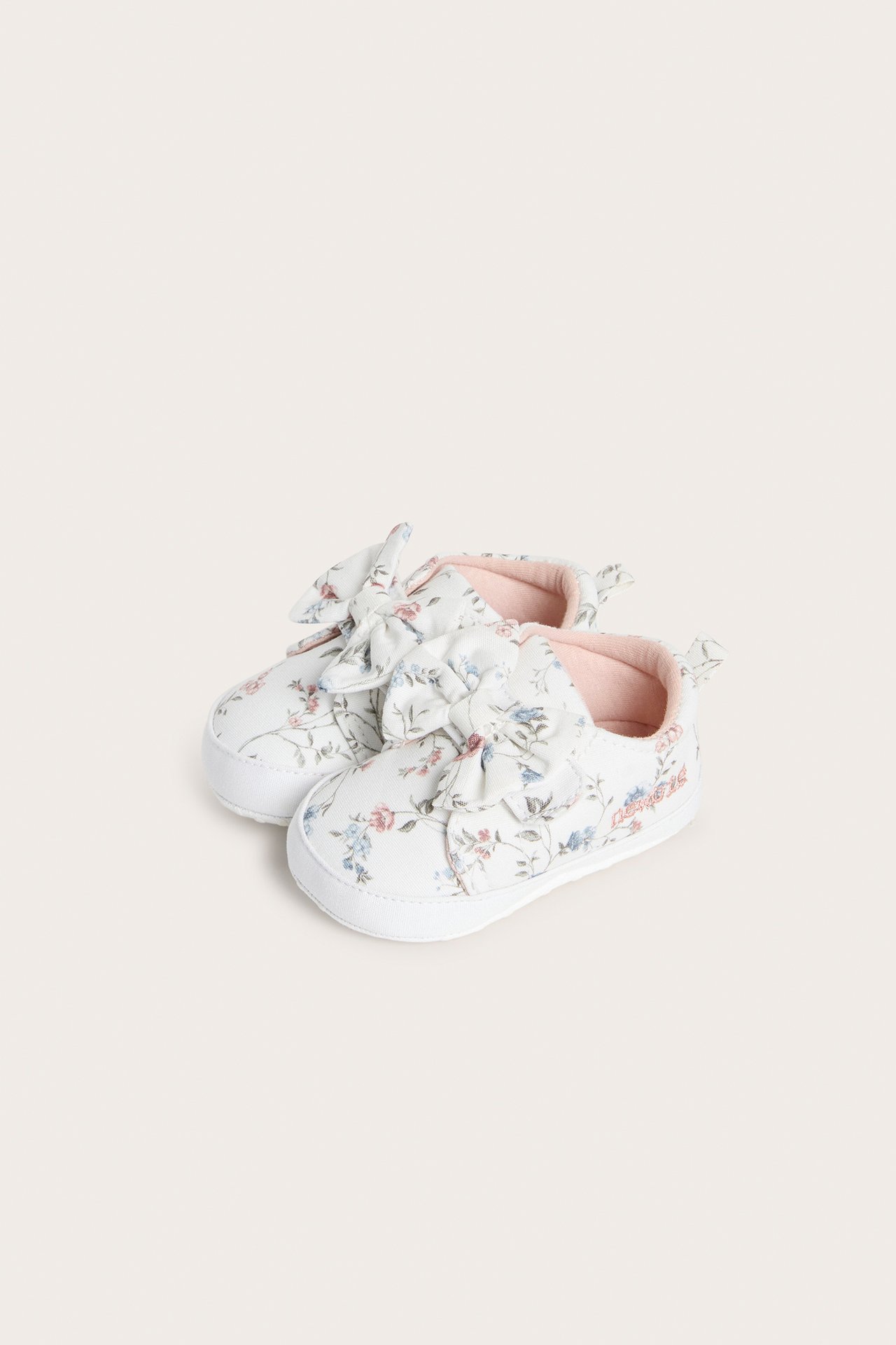 Vorderansicht von cremefarbenen Babyschuhen mit Blumenmuster, großer Schleife und weichem rosa Futter.