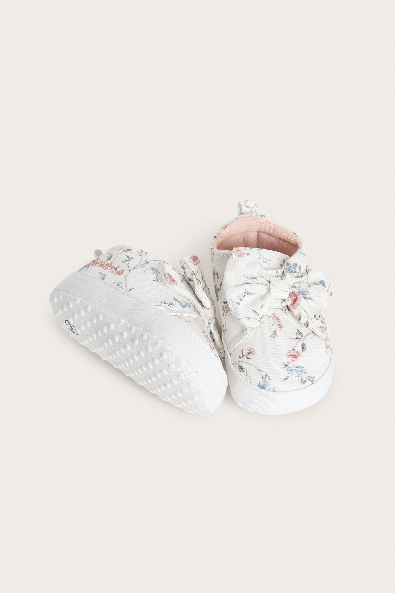 Nahaufnahme von cremefarbenen Babyschuhen mit rosa/blauem Blumenmuster, Zierschleife, Klettverschluss und Antirutschsohle.