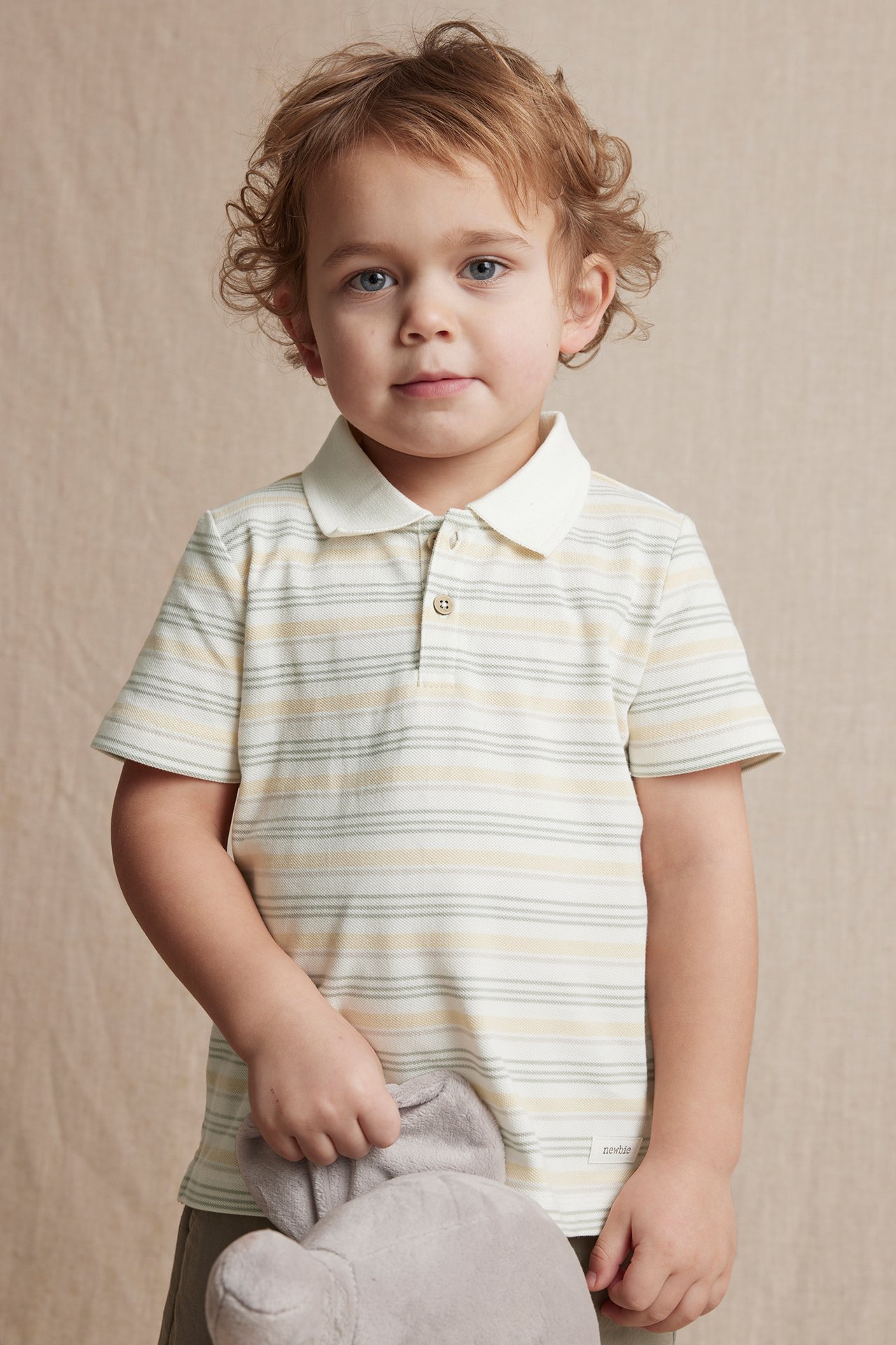 Front view of a kid wearing an off-white striped cotton polo shirt with a collar, holding a grey plush toy.