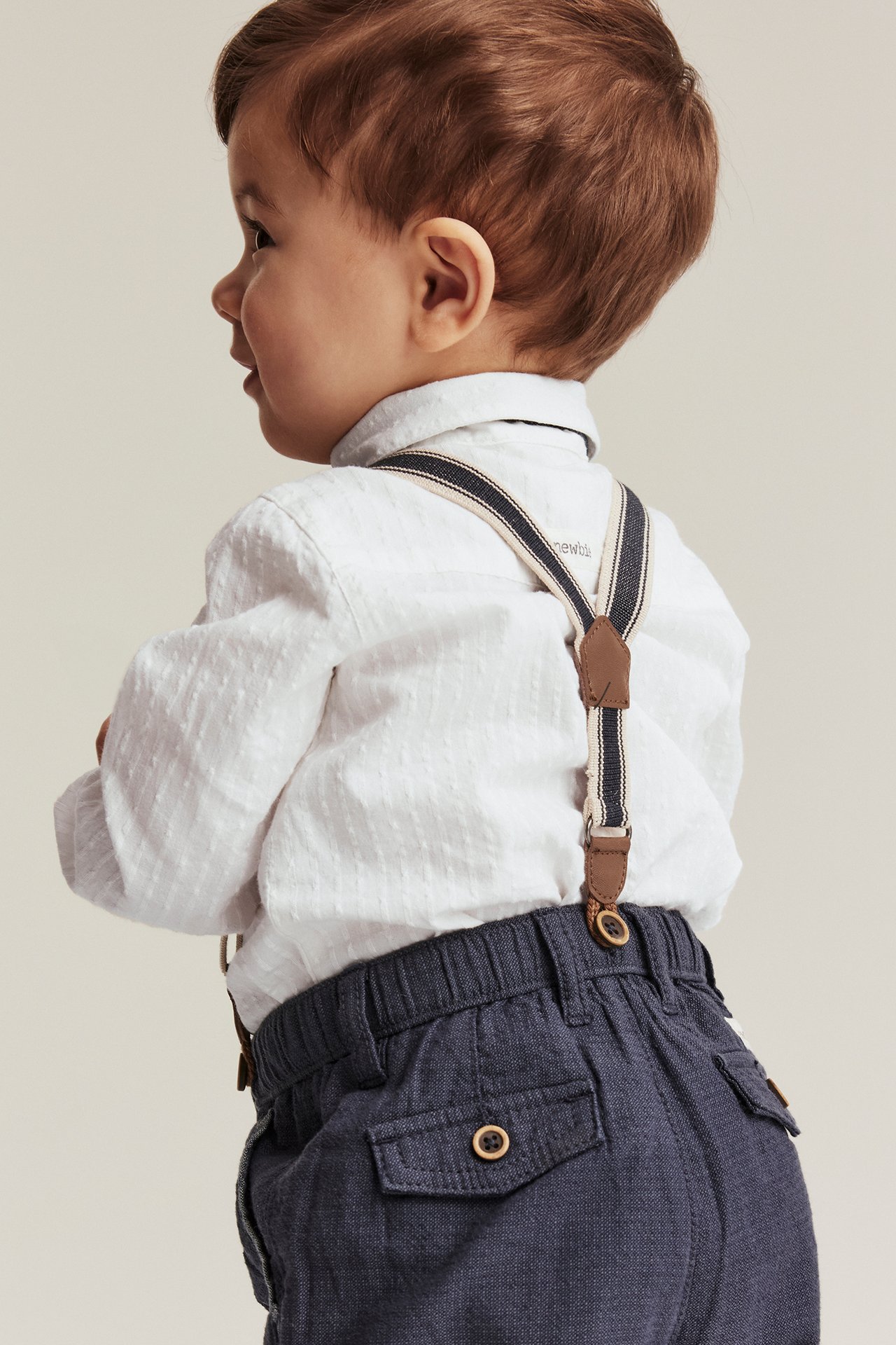 Side view of a baby boy wearing an off-white textured long-sleeved shirt body, dark blue trousers, and striped suspenders.