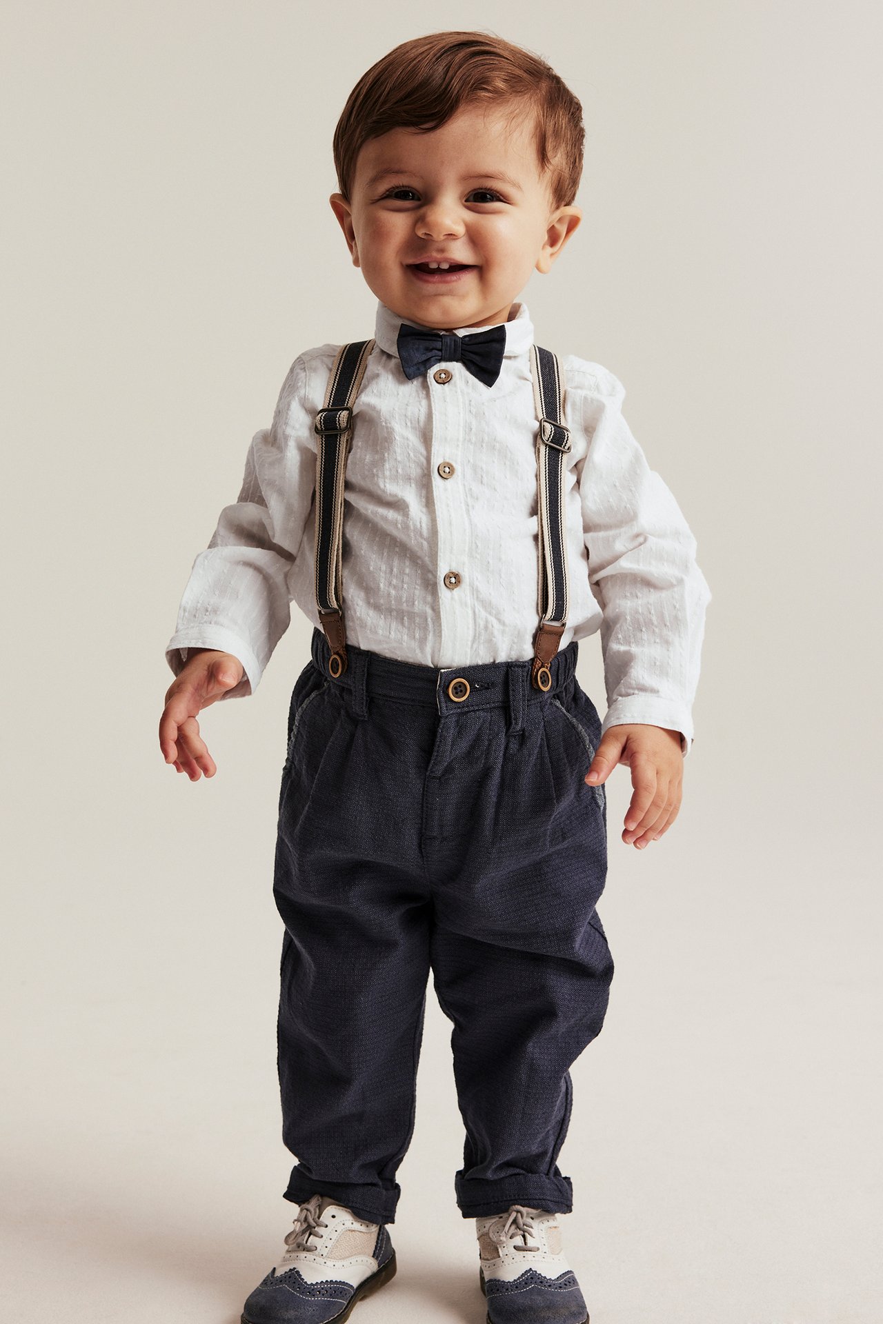Front view: Baby boy wears off-white woven shirt body with dark blue bow tie, dark blue trousers, suspenders.