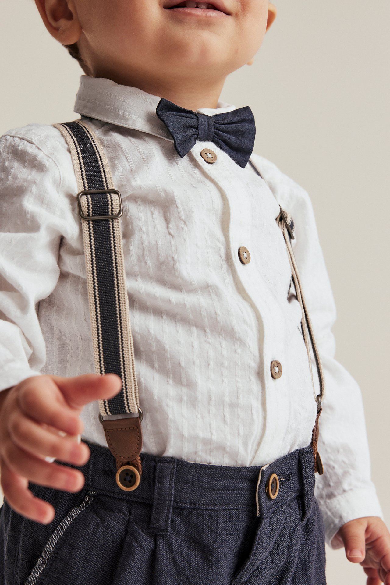 Front view: baby in off-white woven shirt bodysuit, dark blue bow tie, trousers, and striped suspenders.
