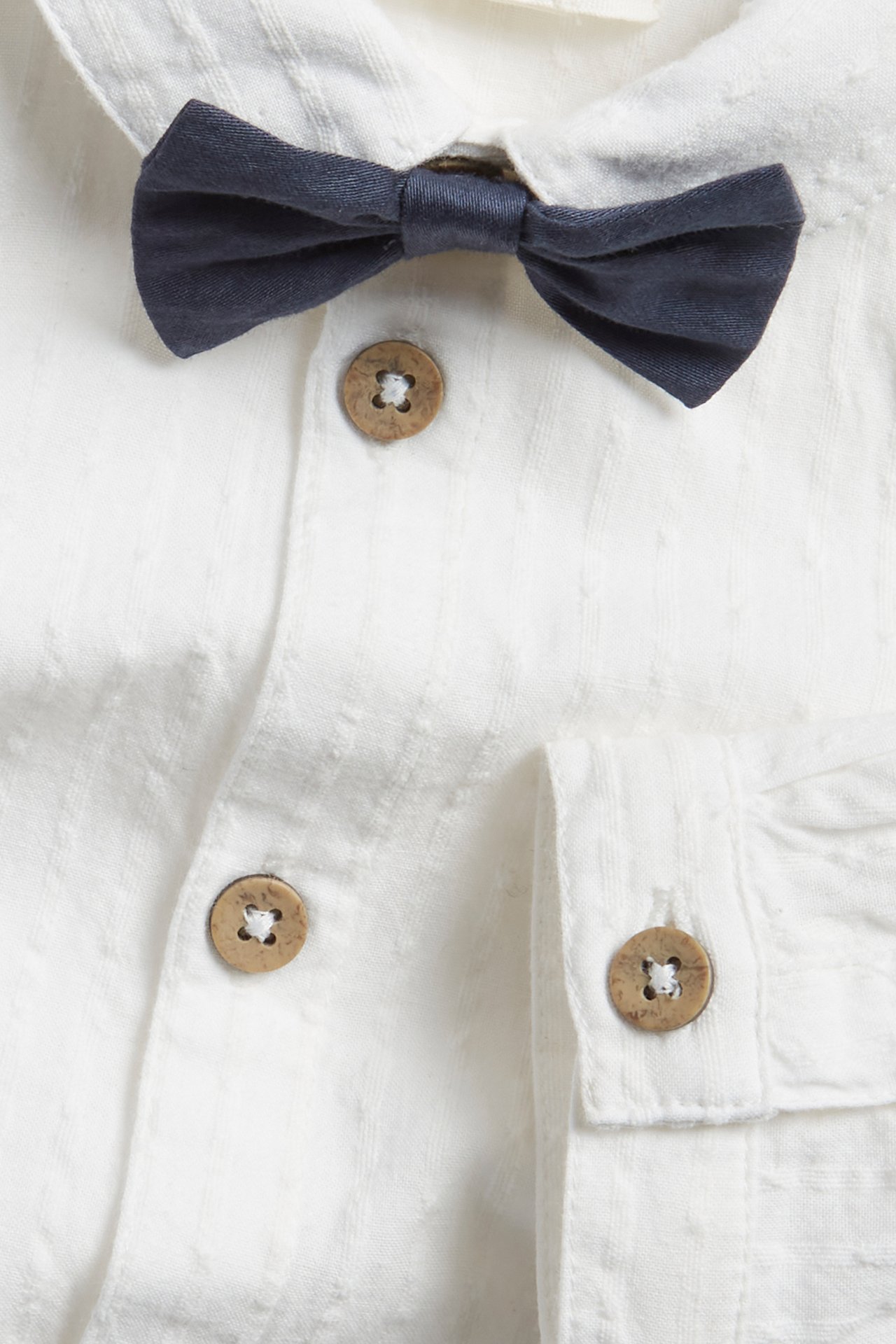 Close-up of a baby's off-white woven cotton shirt-bodysuit with wooden buttons and a dark blue bow tie.