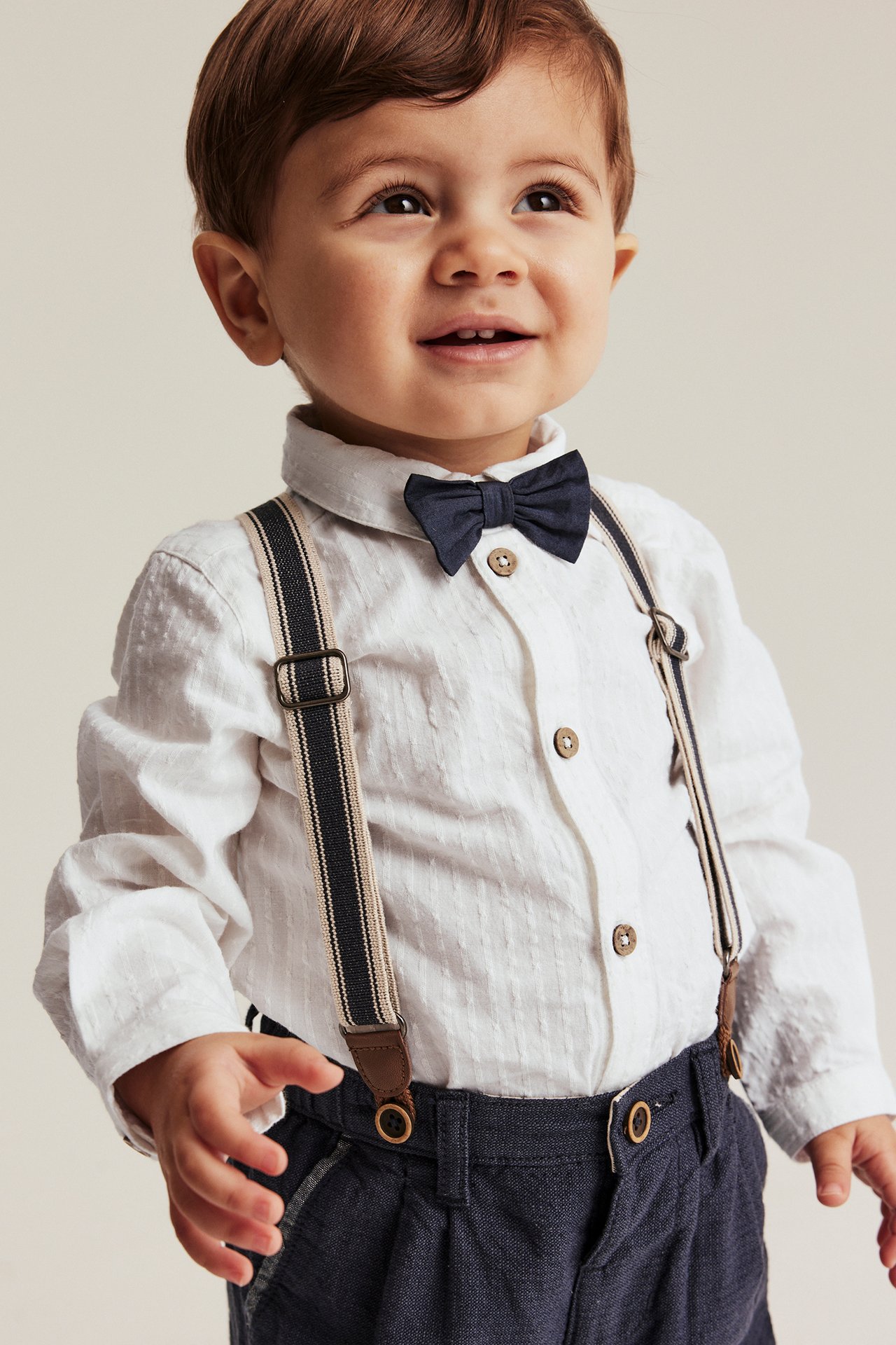 Front view: Baby boy in off-white woven shirt body, dark blue bow tie, trousers, and striped suspenders.