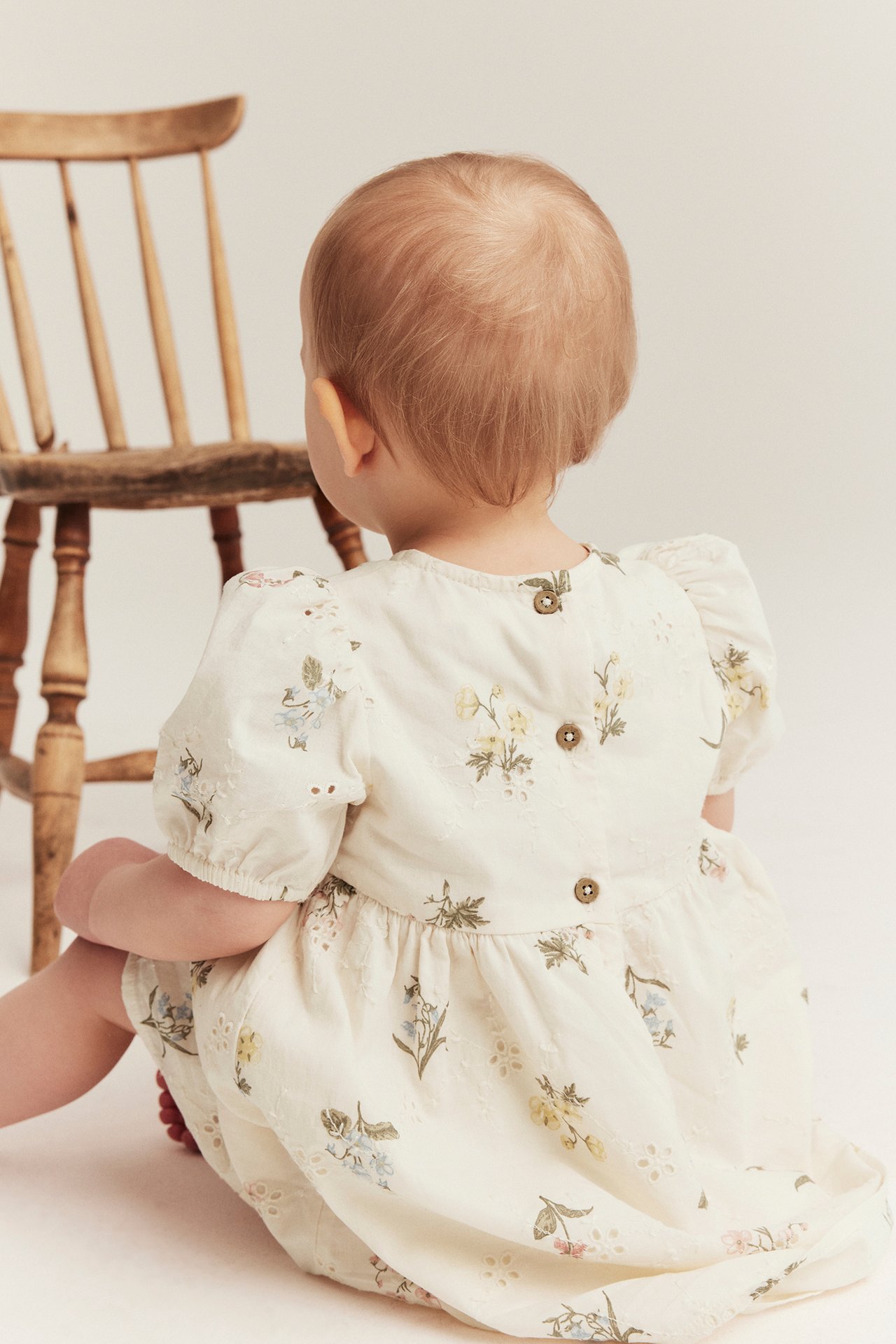 Rückenansicht eines Babys in einem cremefarbenen Broderie Anglaise Kleid mit Blumenmuster und Knöpfen.