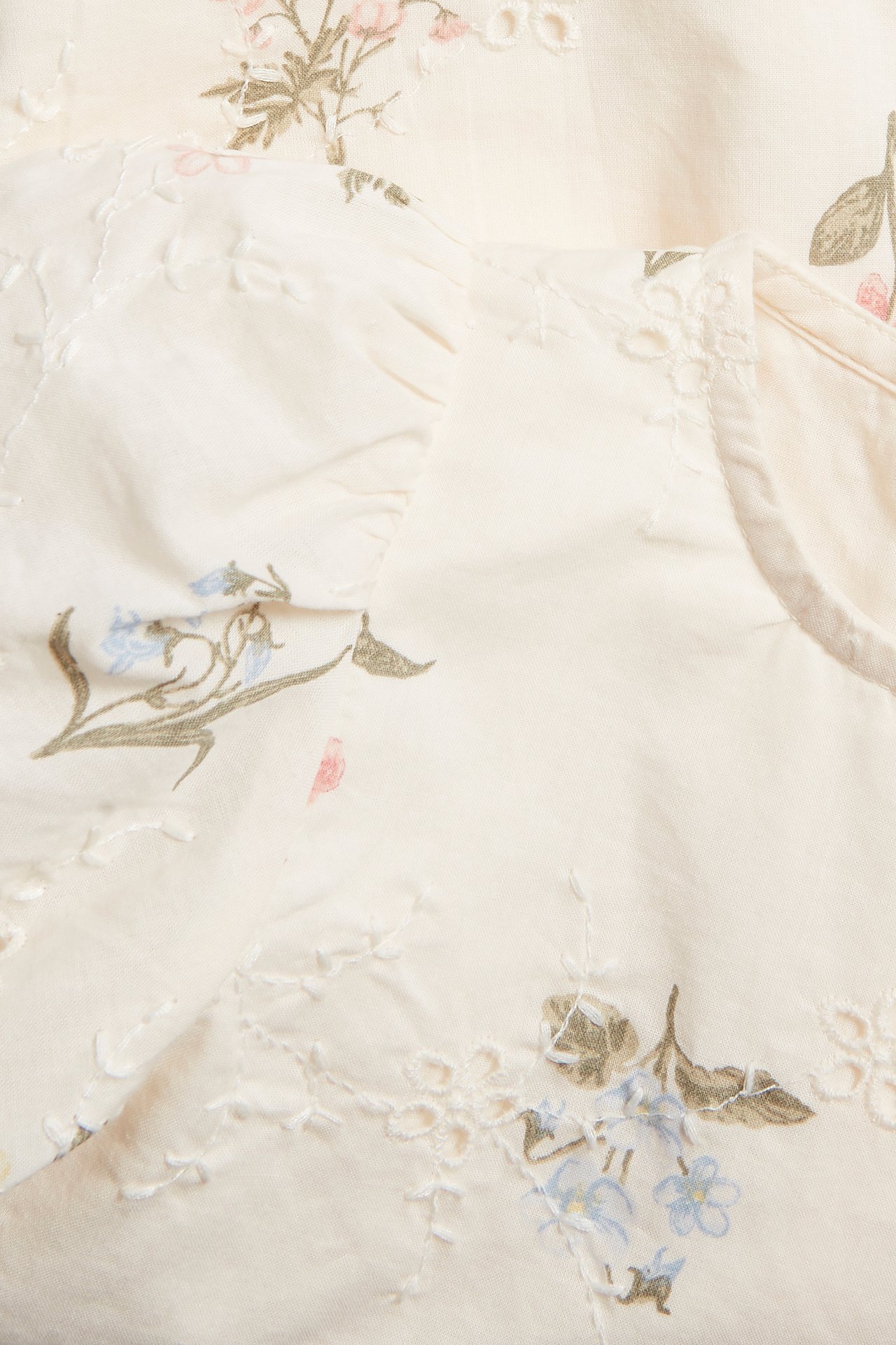 Nahaufnahme eines cremefarbenen gewebten Kinderkleides mit rosa-blauem Blumendruck und Broderie Anglaise Details.