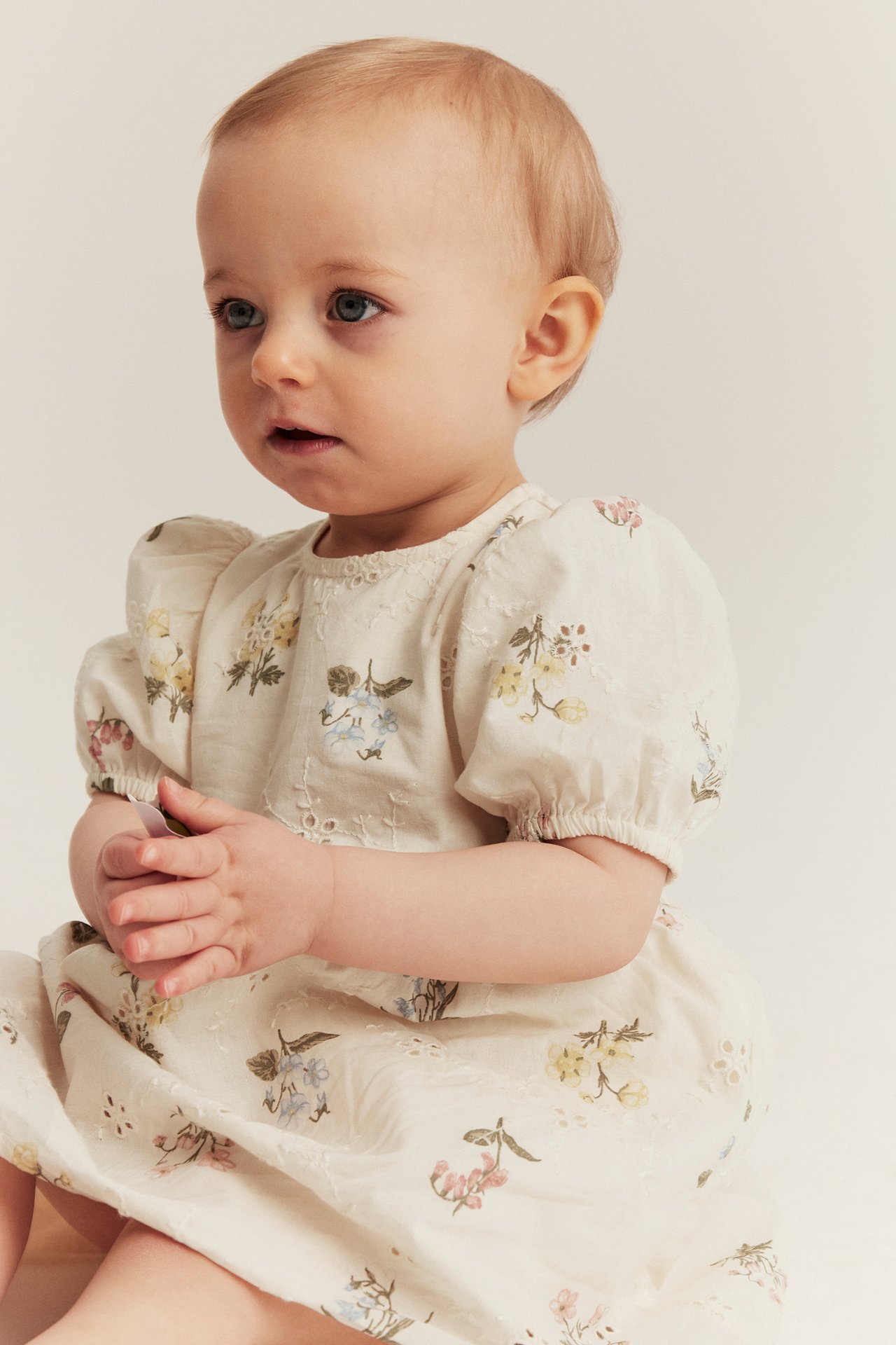 Seitenansicht eines Babys in einem cremefarbenen Broderie Anglaise Kleid mit Blumenmuster und Puffärmeln.