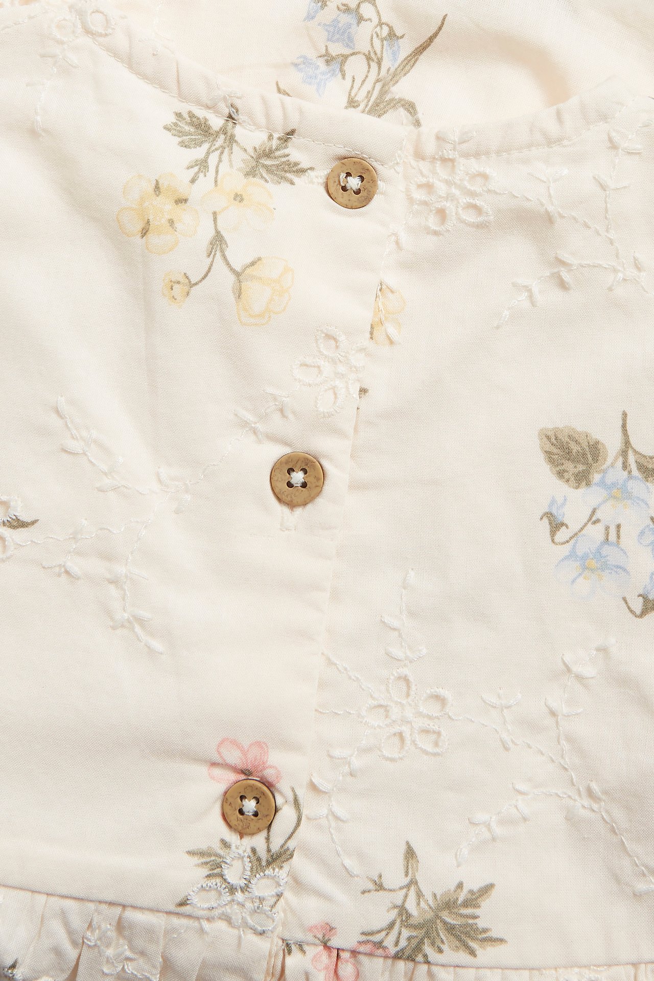 Nahaufnahme eines cremefarbenen Kinderkleides mit gedrucktem Blumenmuster, Broderie Anglaise und Holzknöpfen.