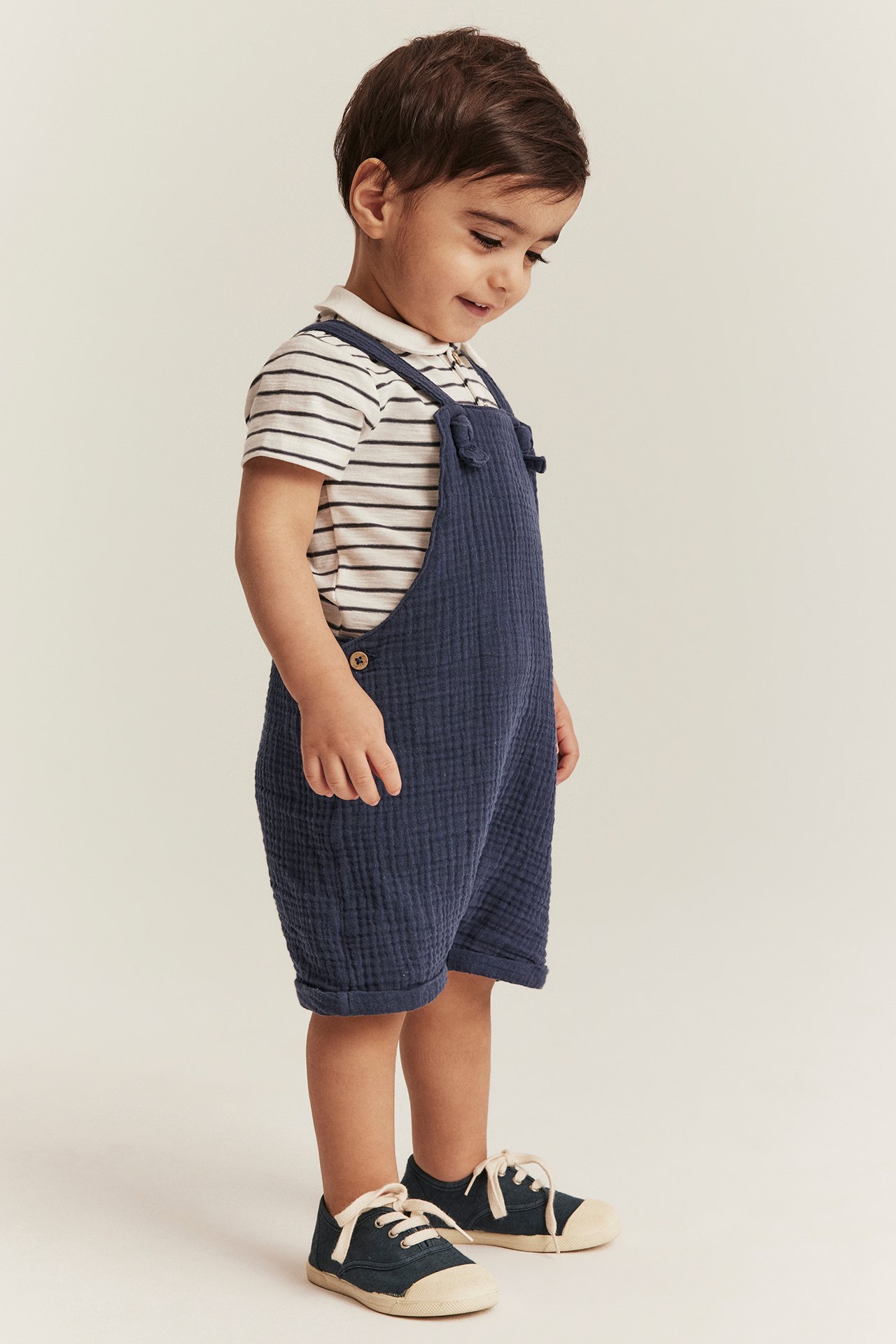 Side view of a kid wearing dark blue woven dungaree shorts over a striped polo shirt and canvas shoes.