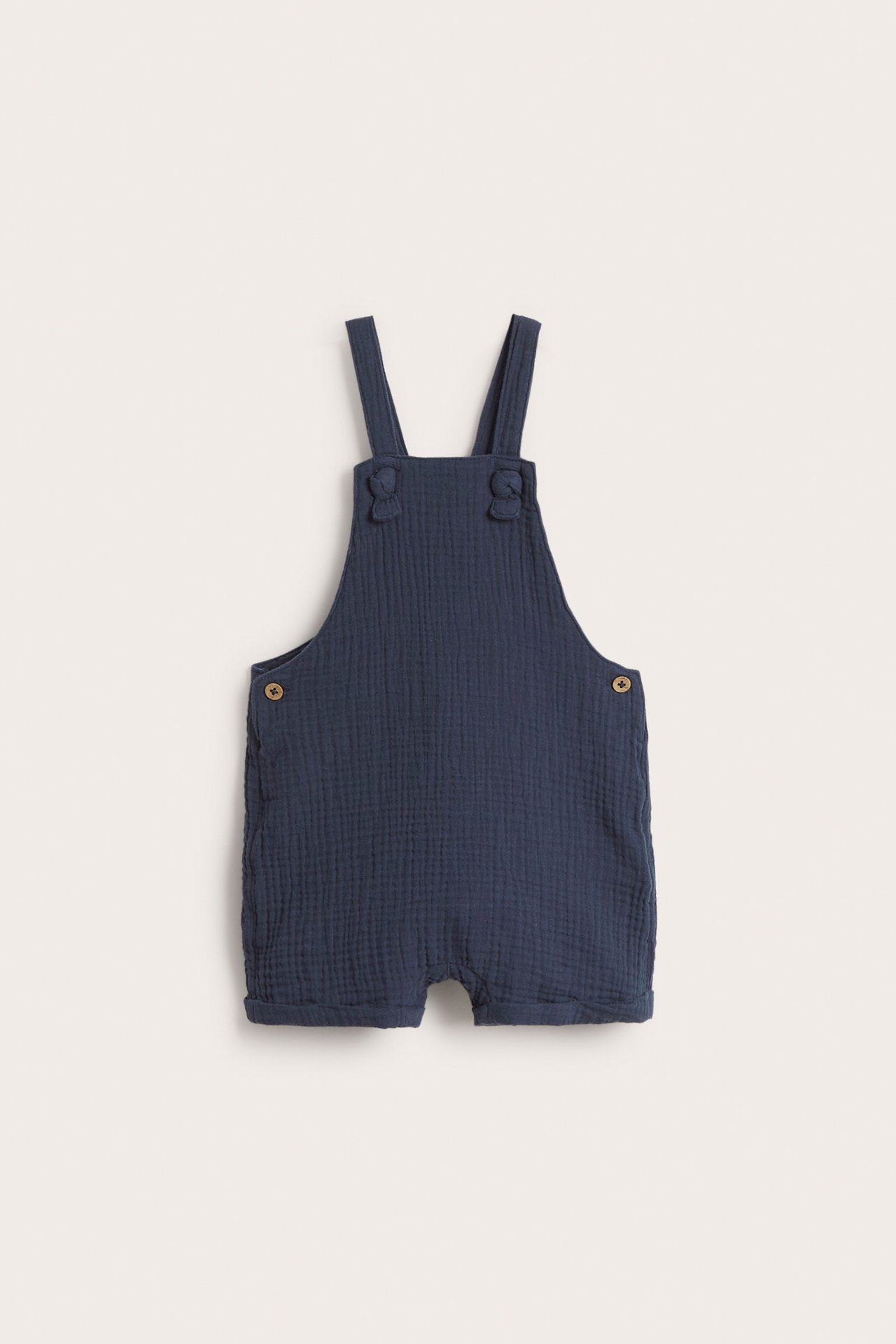 Front view of dark blue woven cotton baby dungaree shorts with adjustable knotted straps and side buttons.