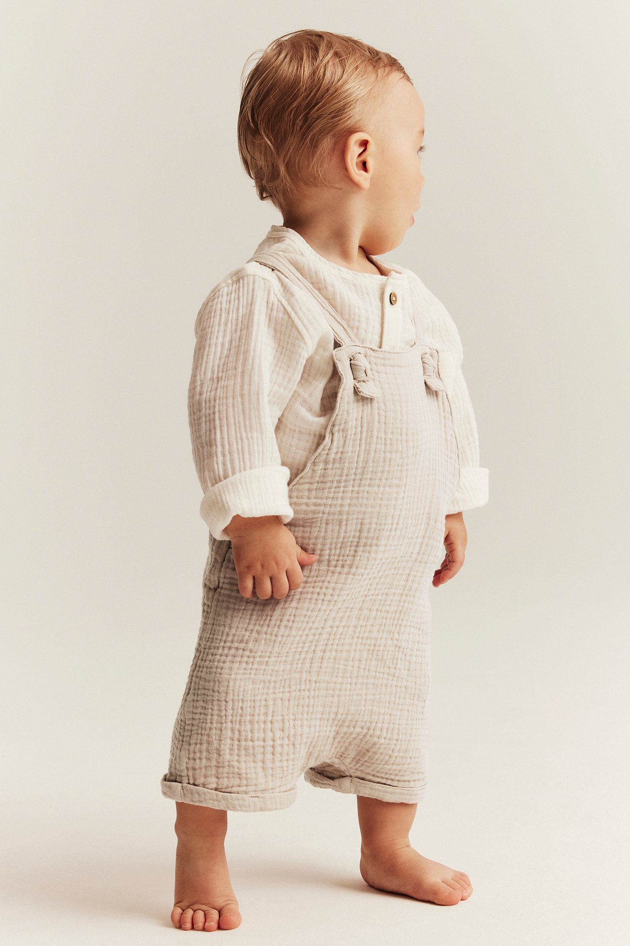 Seitenansicht eines Babys in beigen gewebten Latzshorts mit geknoteten Trägern über einem hellen Langarmshirt.