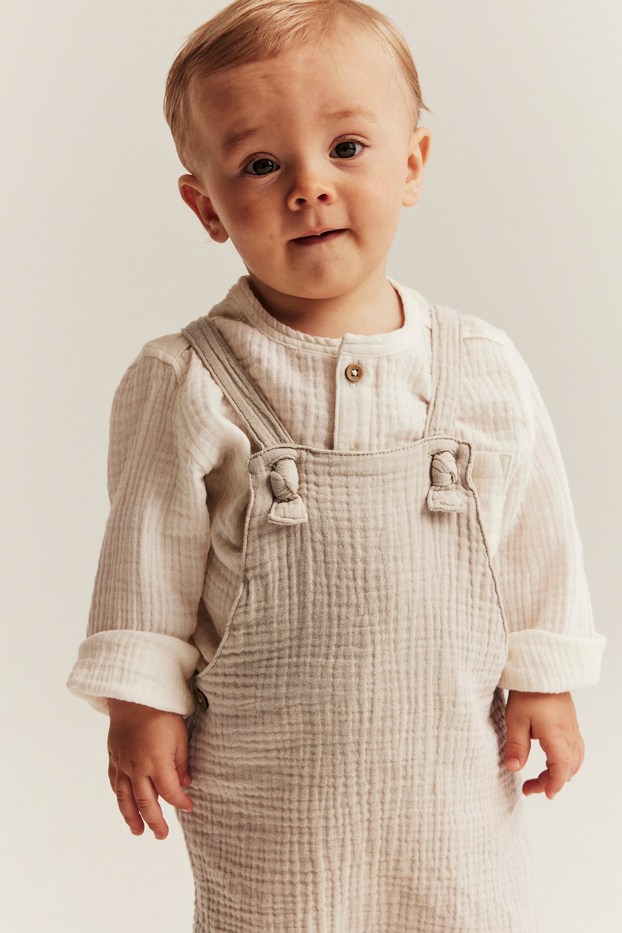 Vorderansicht eines Babys in beigen gewebten Baumwoll-Latzshorts mit geknoteten Trägern über einem weißen Langarmshirt.