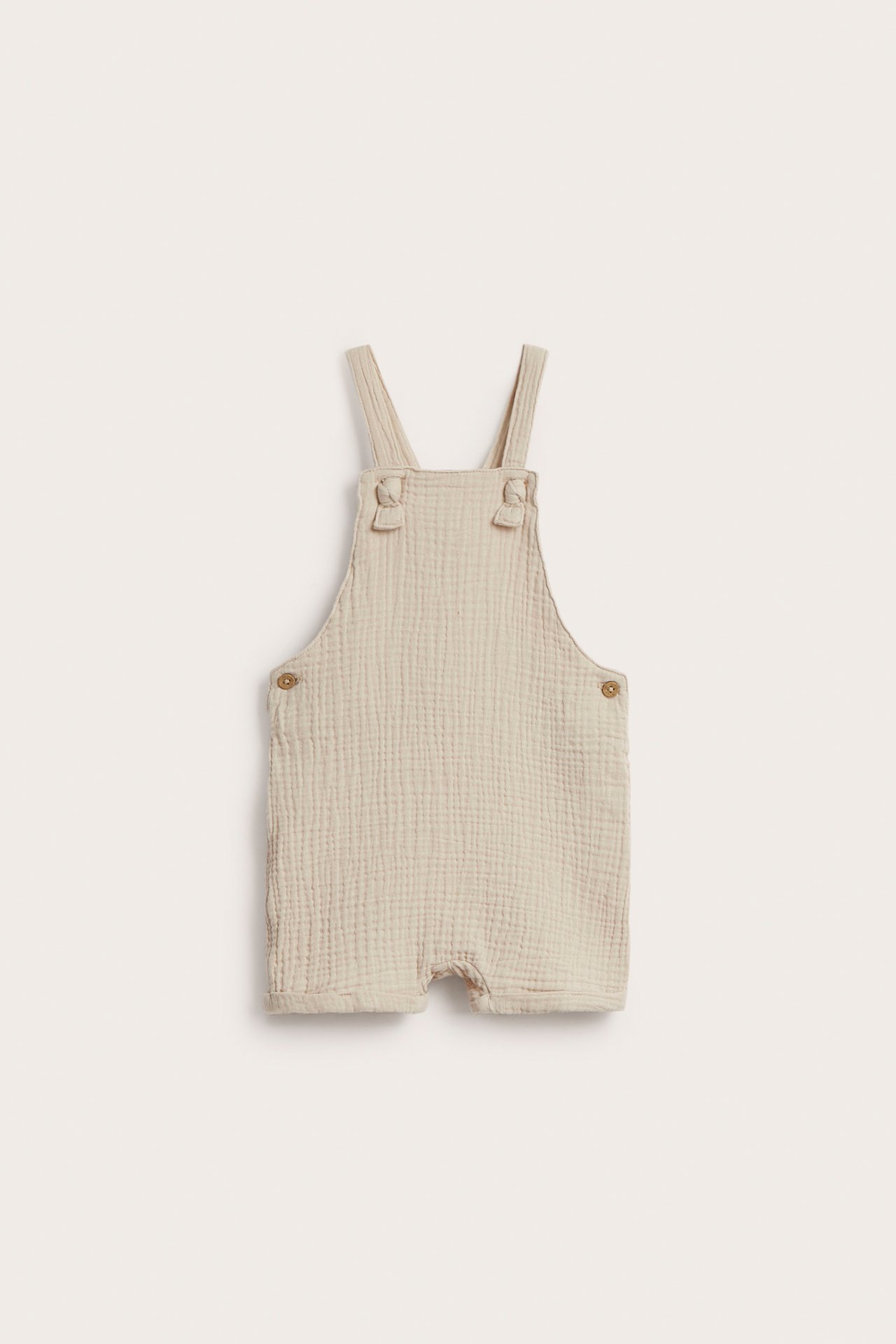 Vorderansicht beiger gewebter Baumwoll-Baby-Latzshorts mit verstellbaren geknoteten Trägern und Seitenknöpfen.