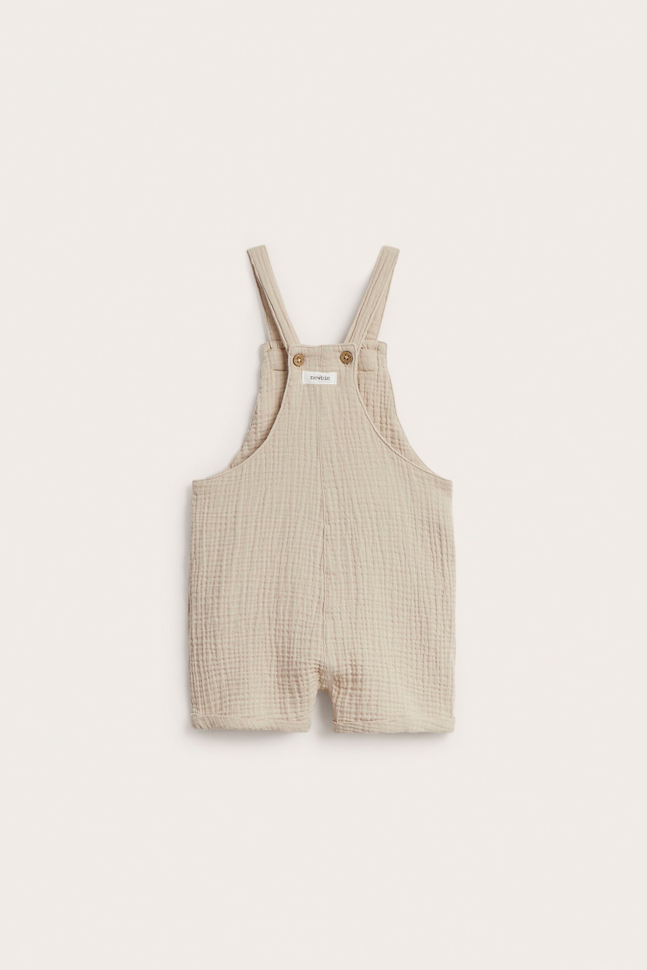 Vorderansicht beiger gewebter Baumwoll-Baby-Latzshorts mit verstellbaren Knöpfen an den Trägern und Seitenöffnungen.