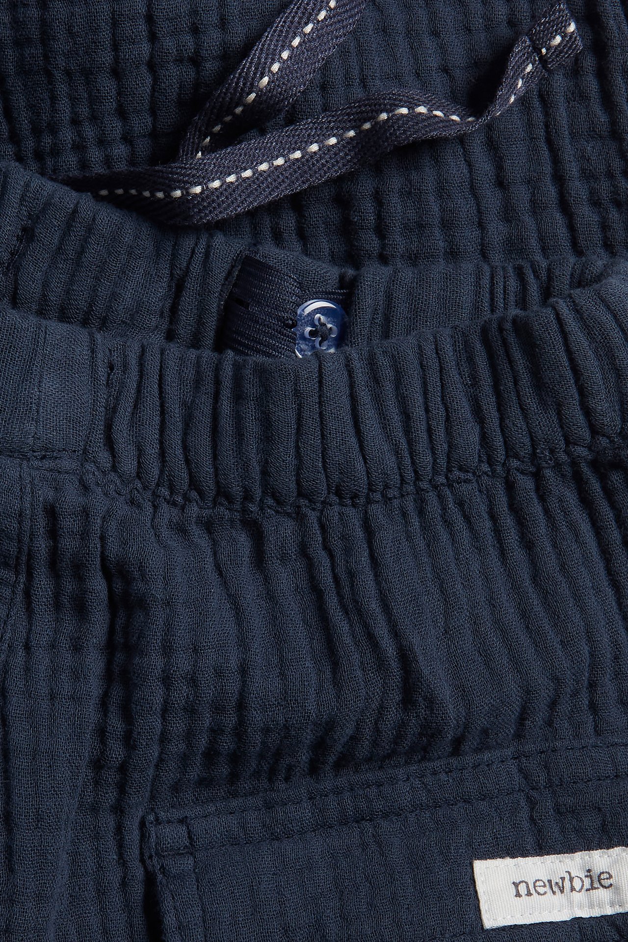 Close-up of dark blue woven cotton pants with elasticated waist, herringbone drawstring, and button.