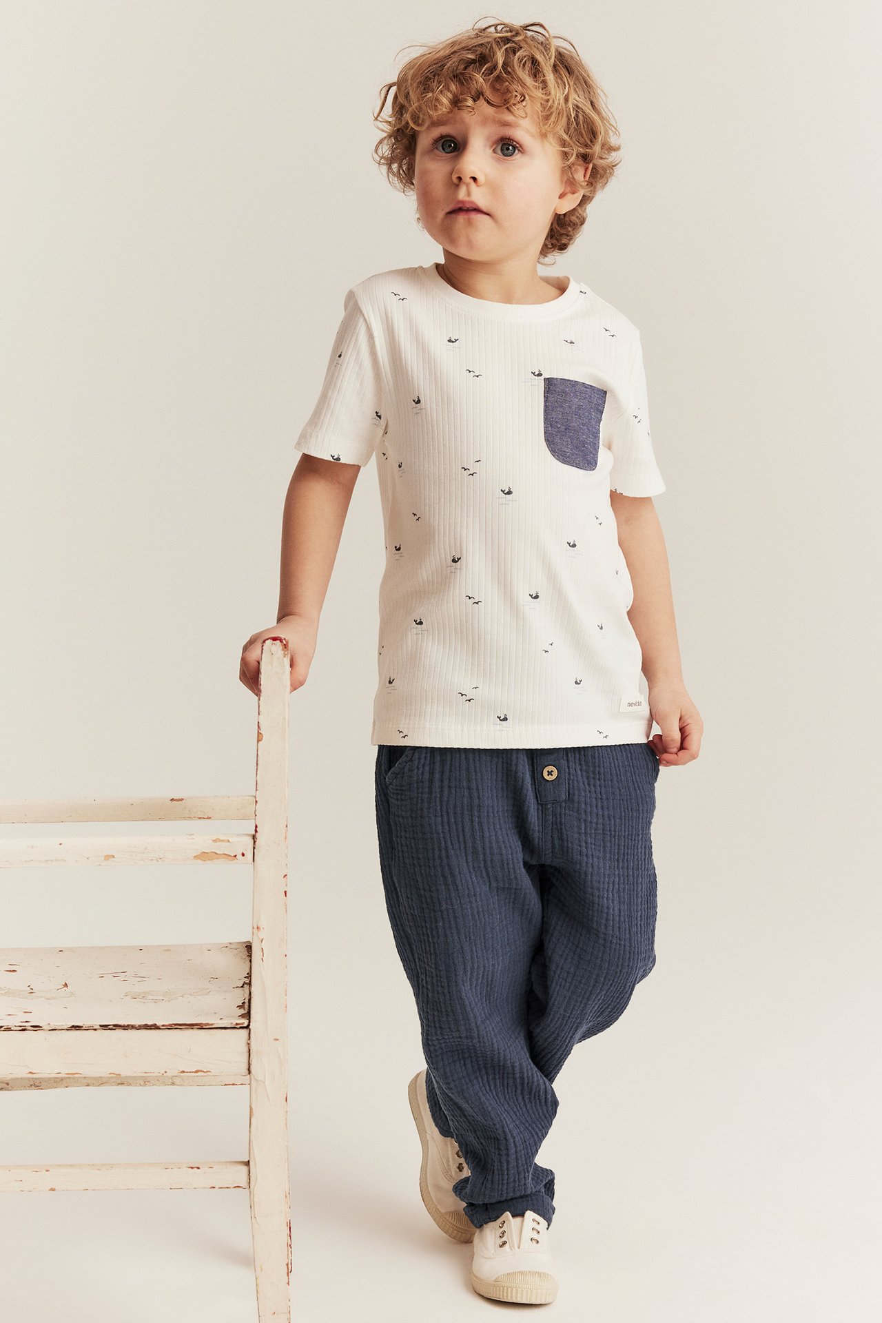 Front view: Kid in dark blue crinkled cotton trousers with front button, and a white ribbed t-shirt.