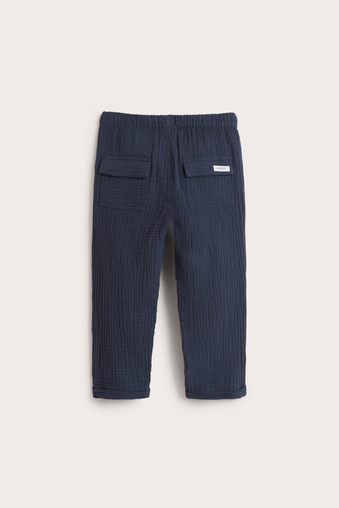 Back view of dark blue woven cotton kid's pants with elastic waist, two flap back pockets, and rolled-up cuffs.