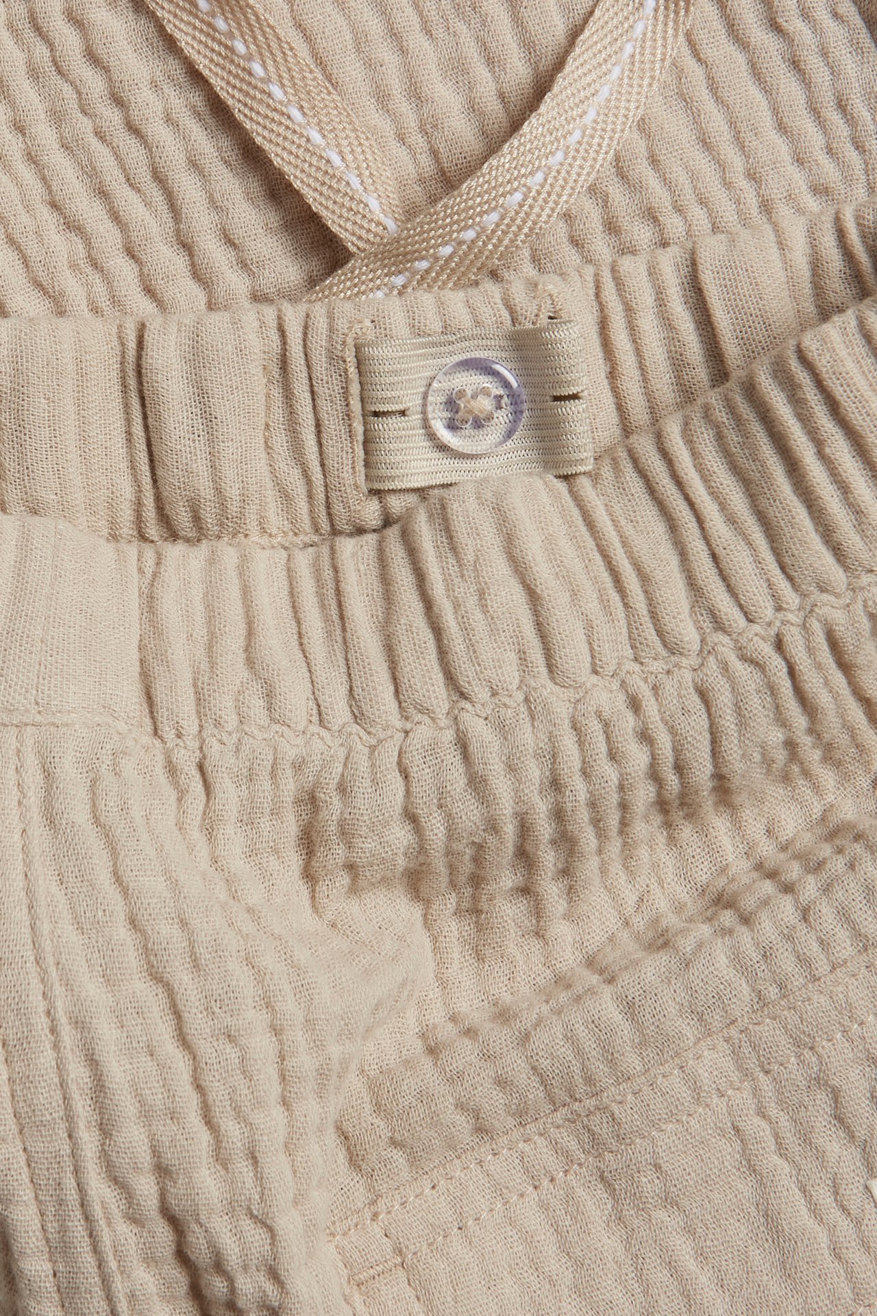 Close-up of beige woven kid's trousers with an elasticated waist, adjustable button, and herringbone drawstring.