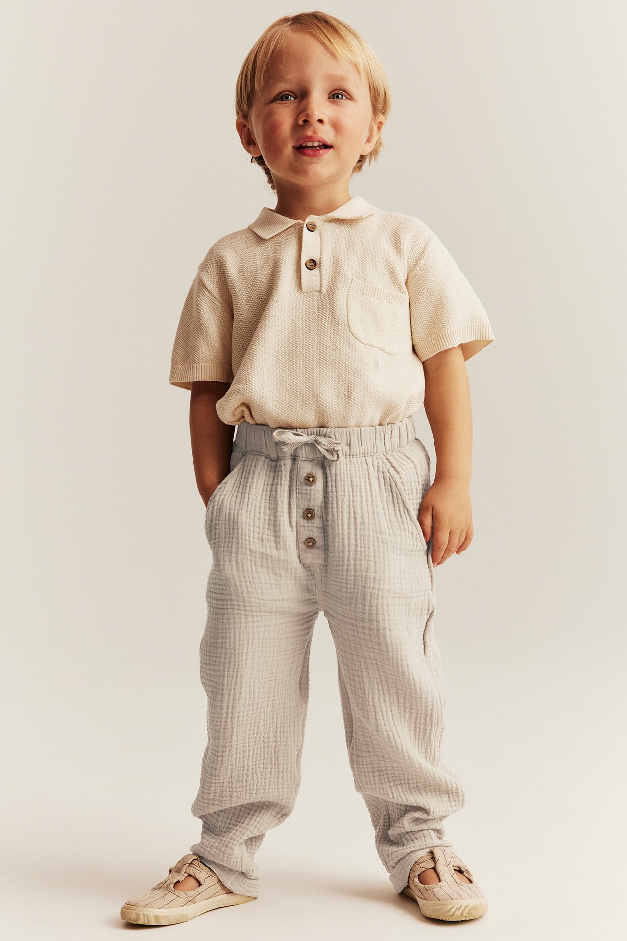 Front view of a kid in light beige woven pants with buttons, a light beige polo shirt, and beige shoes.