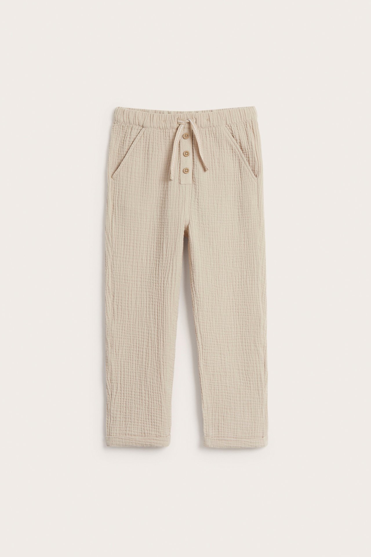 Front view of beige woven cotton pants with elastic waist, drawstring, decorative buttons, and front pockets.
