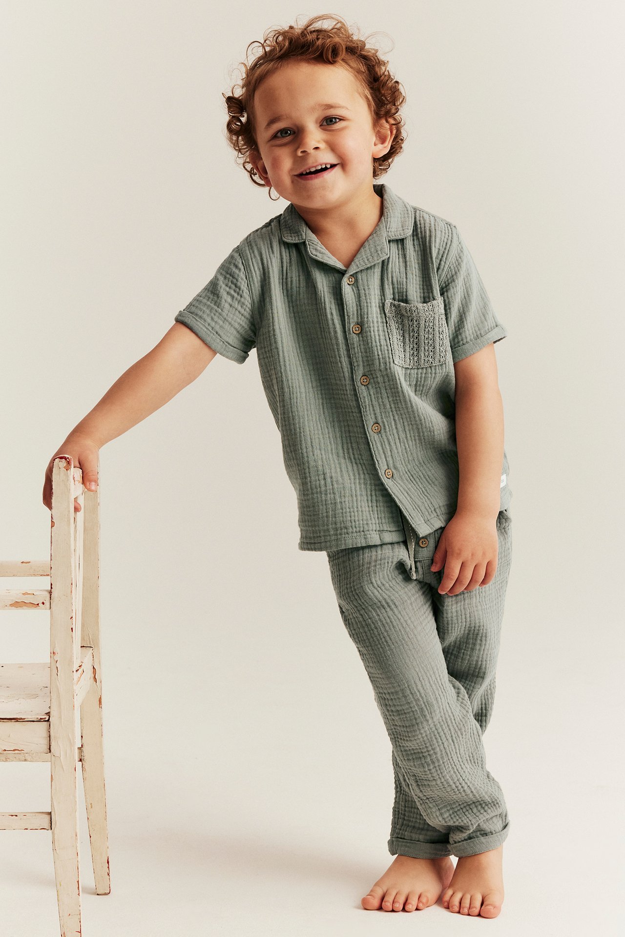 Front view: Smiling kid in green woven pants with rolled cuffs and matching short-sleeved shirt, leaning on chair.