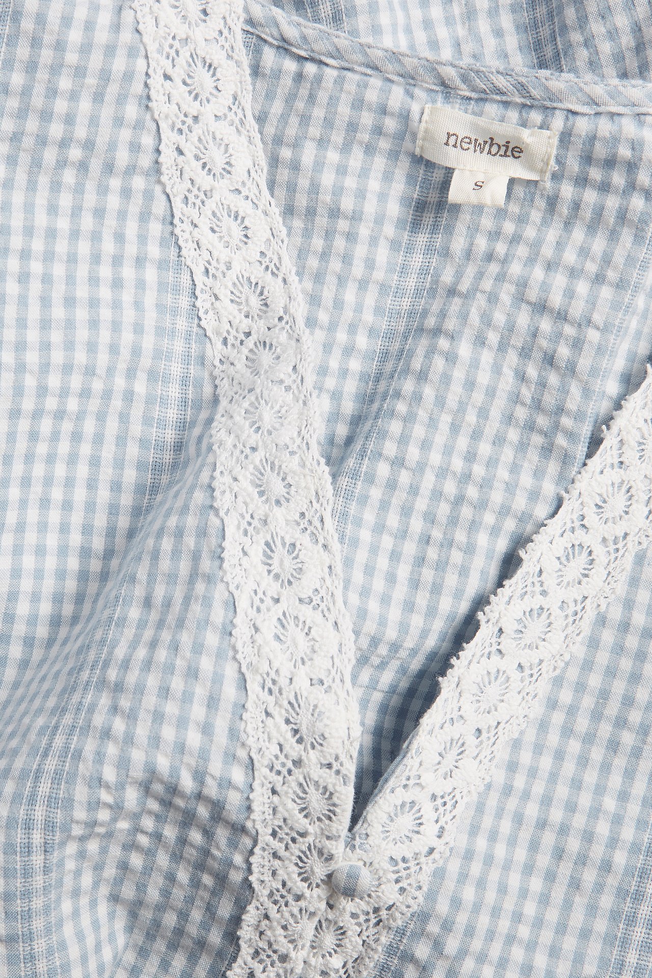 Close-up: Light blue and white gingham cotton blouse, V-neck with white lace trim, covered button.