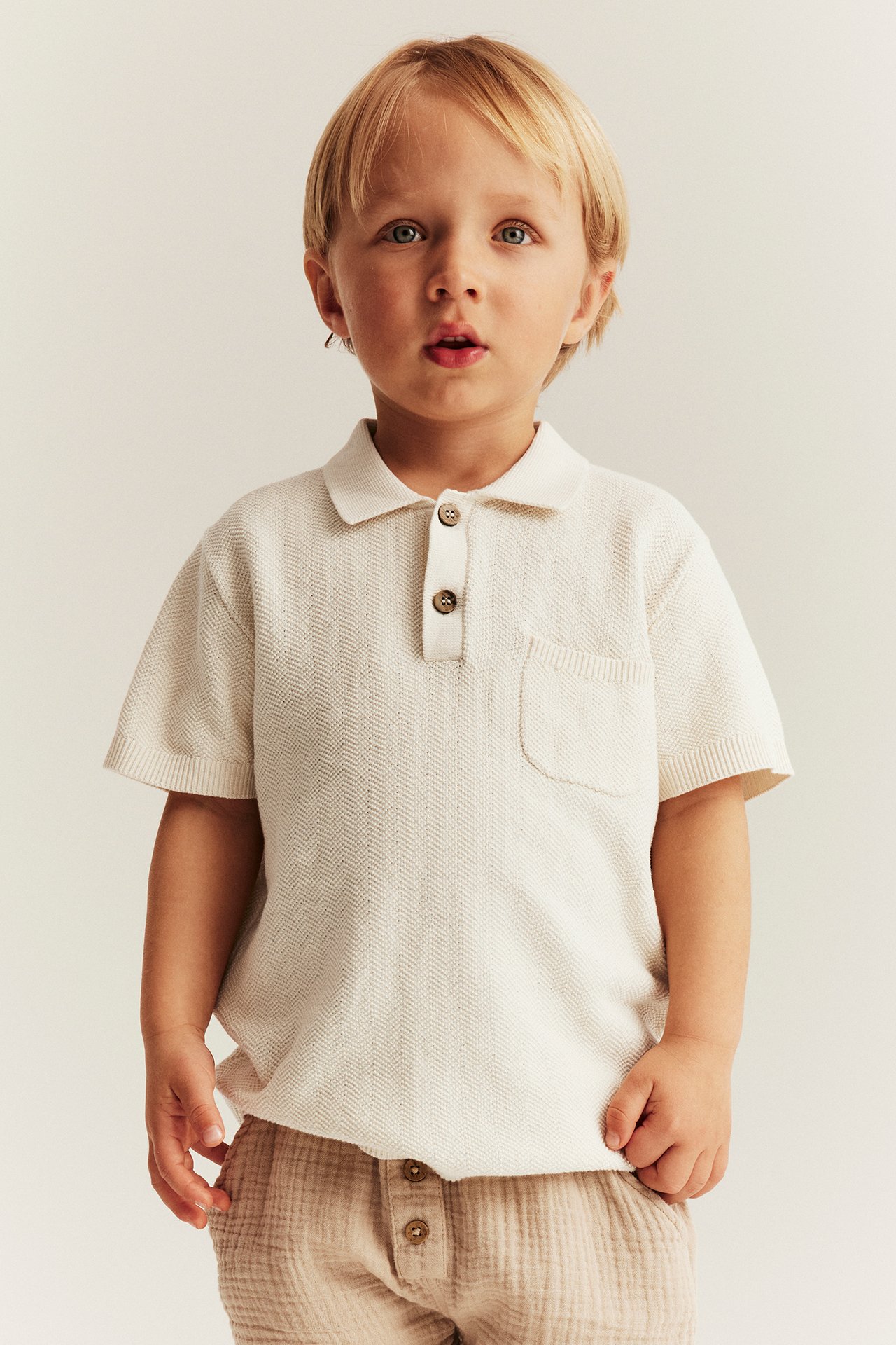 Vorderansicht: Kind in hellbeigem Strick-Poloshirt mit Kragen, Knöpfen, Tasche und beigen Shorts.