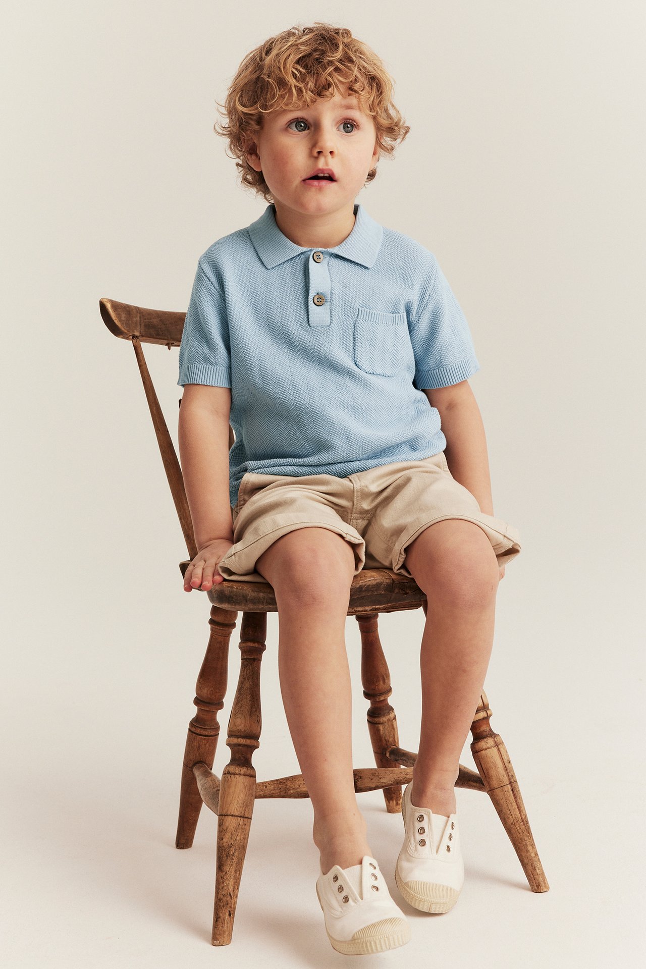Front view of a kid in a light blue knitted polo shirt, beige shorts, and white canvas shoes.