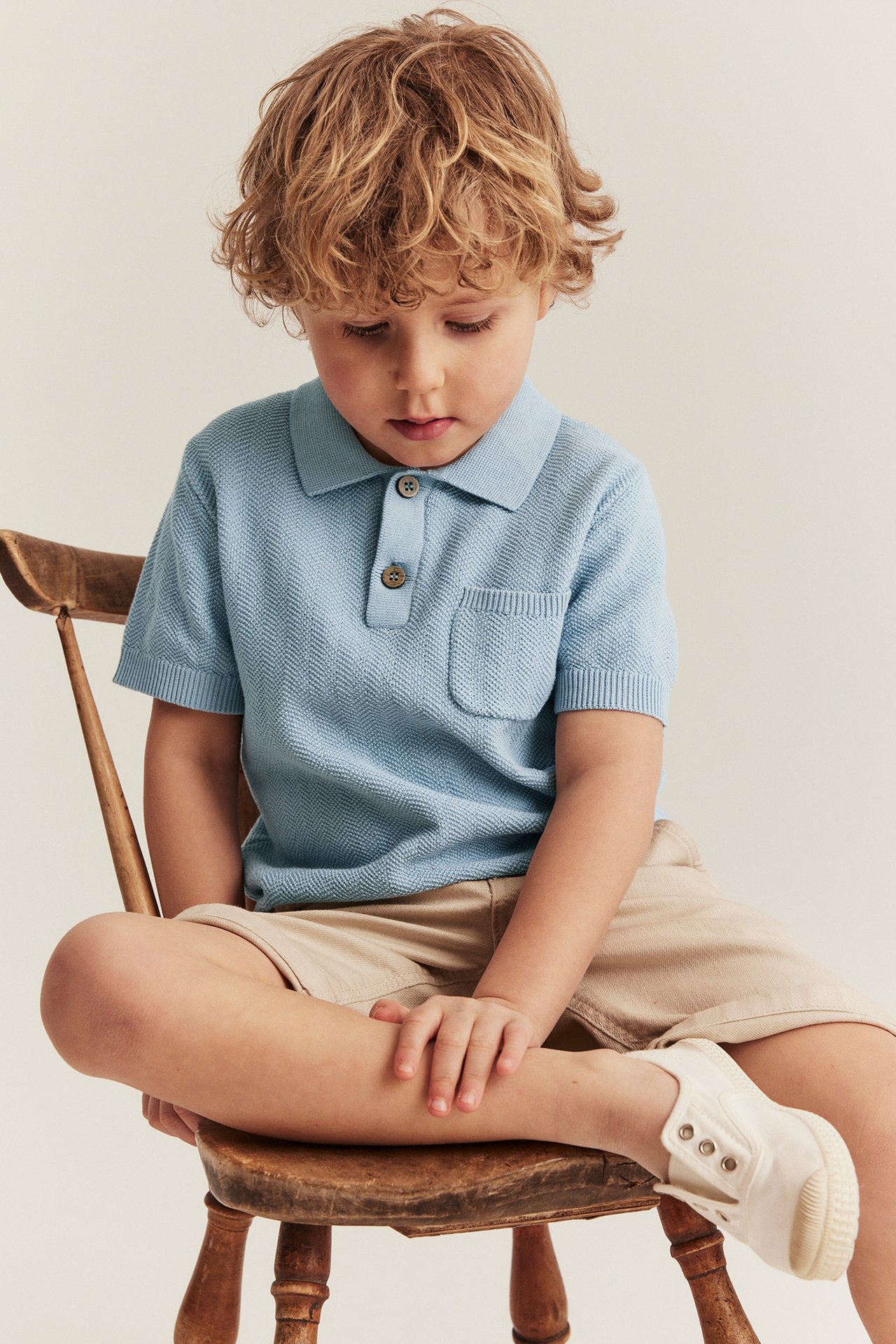 Front view of a kid wearing a light blue knitted polo shirt with buttons and pocket, beige shorts, and white canvas shoes.