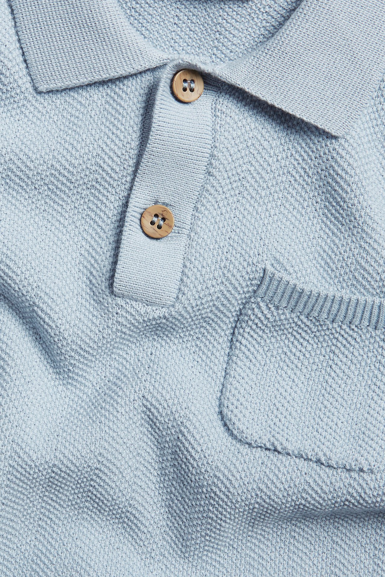 Close-up front view: light blue cotton knitted polo shirt with collar, two wooden buttons, and chest pocket.