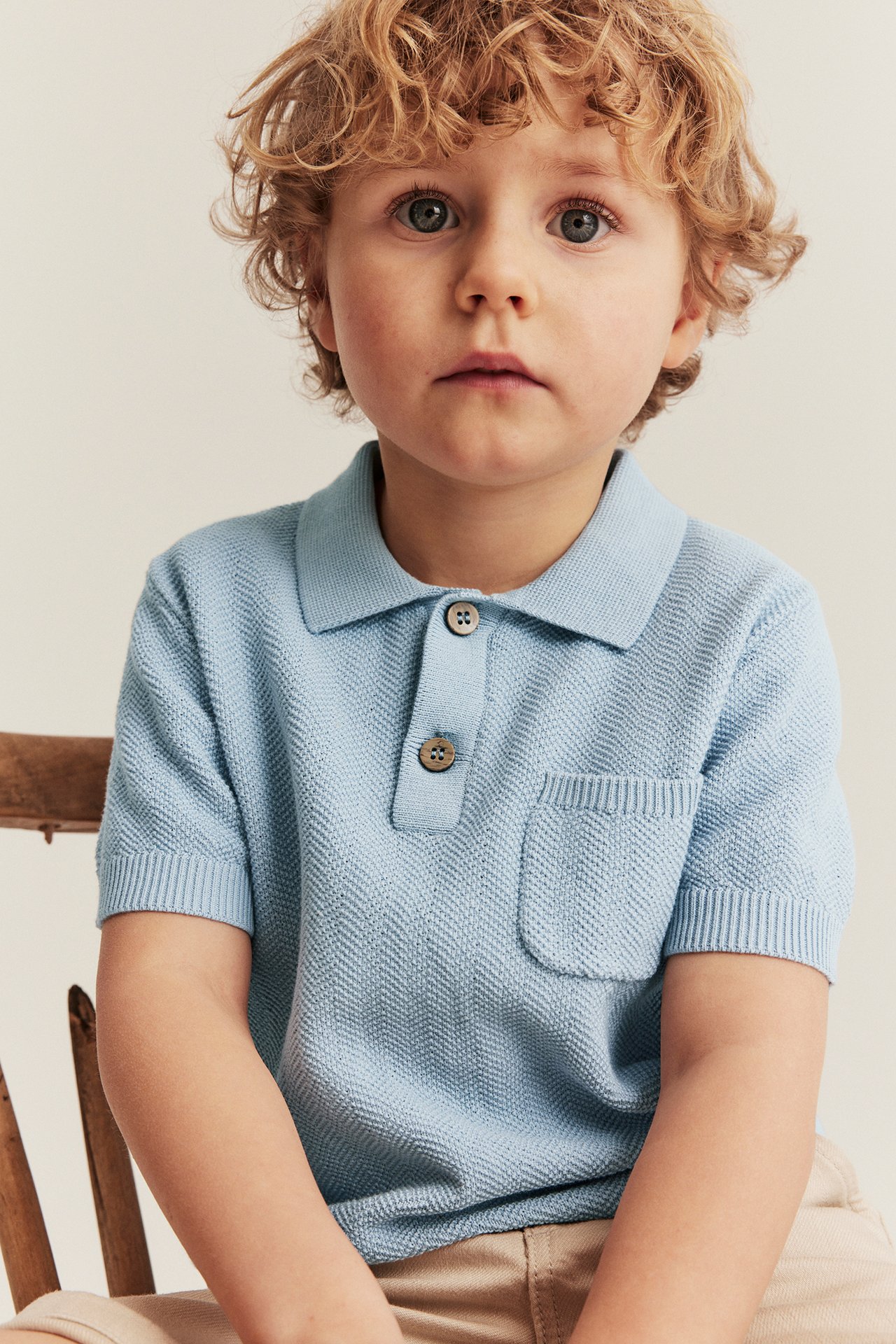Front view of a kid in a light blue knitted polo shirt with collar, buttons, and a pocket.