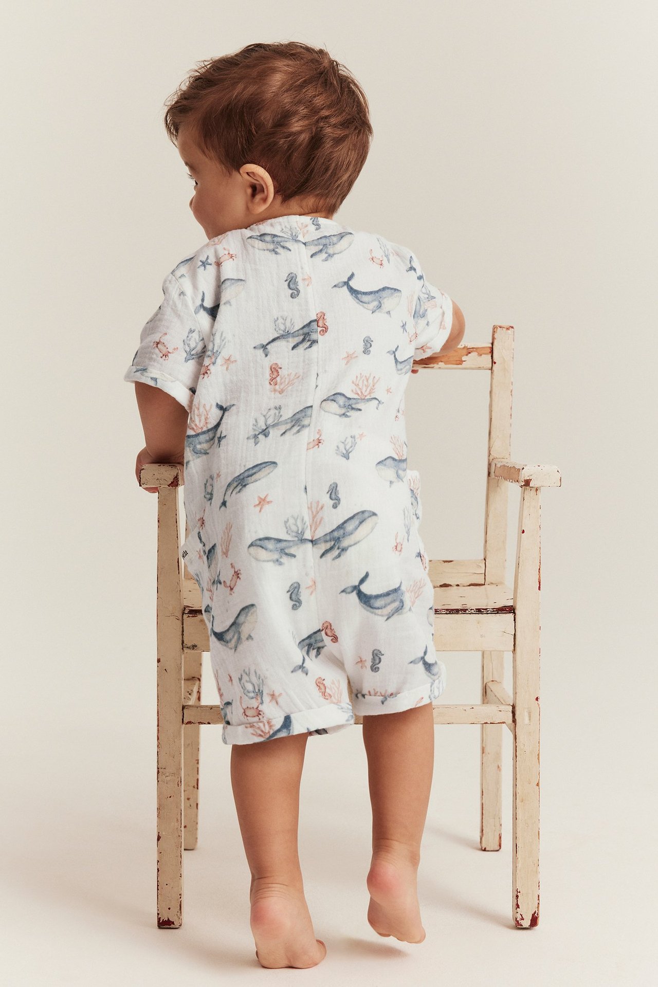 Back view of a baby in an off-white cotton muslin romper with whale print, holding a chair.