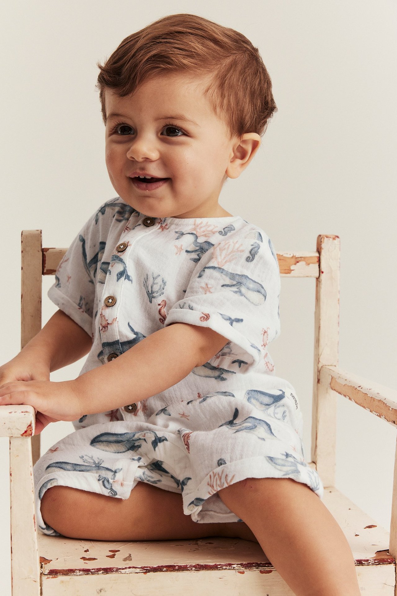 Front-side view of a baby in an off-white cotton muslin romper with sea animal pattern, front buttons.