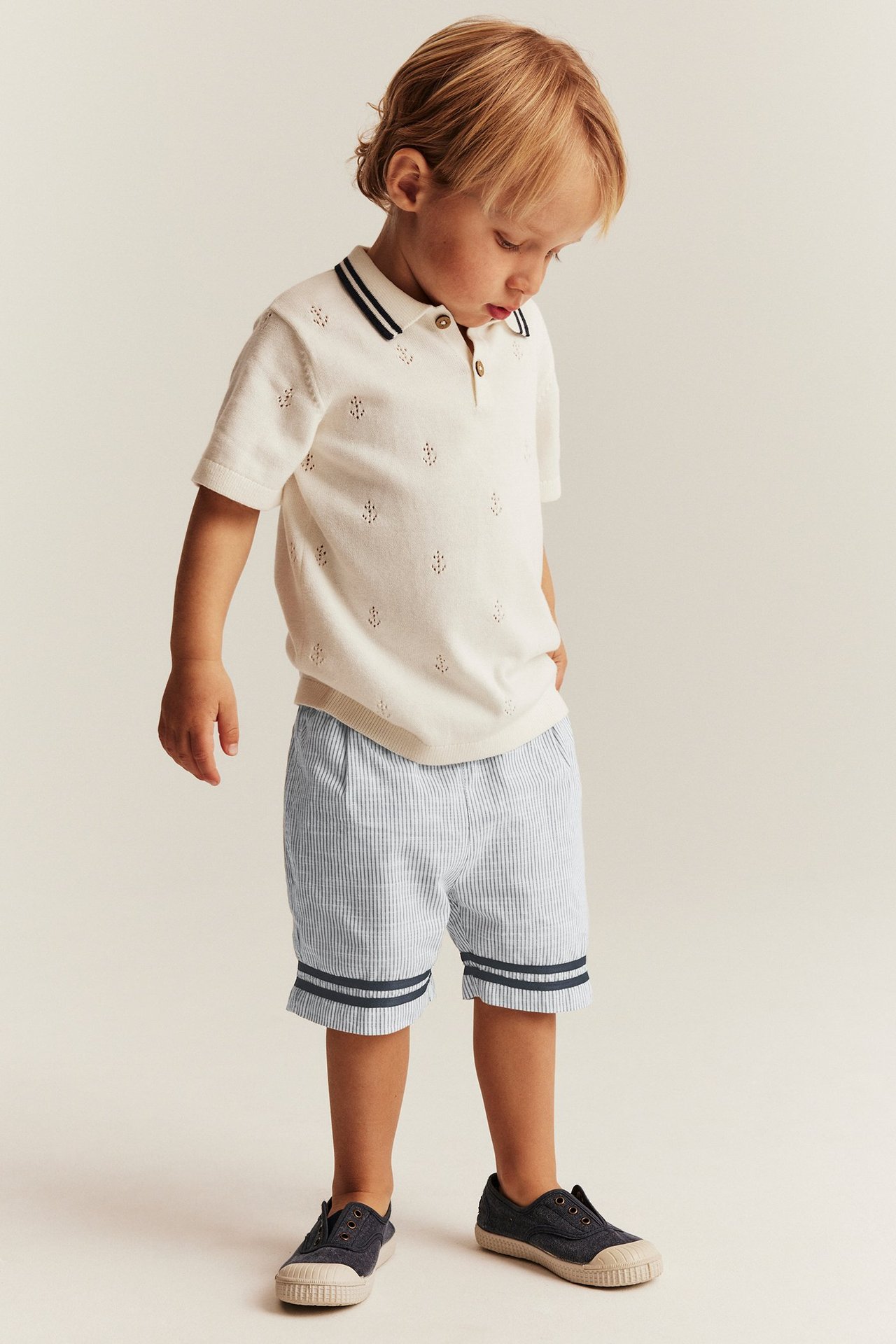 Kind in hellblau gestreiften Shorts mit Knöpfen, cremefarbenem Poloshirt und dunkelblauen Leinenschuhen. Vorderansicht.