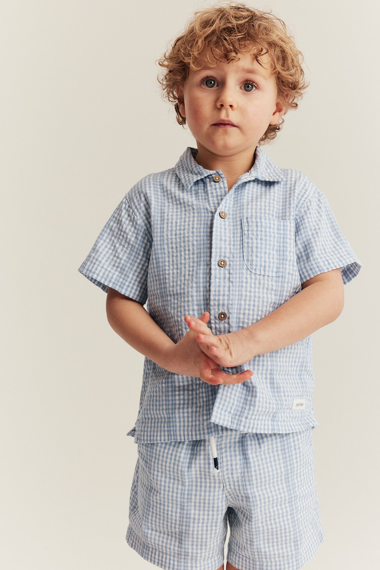 Front view of a kid in light blue and white checked cotton shorts with drawstring and matching shirt.