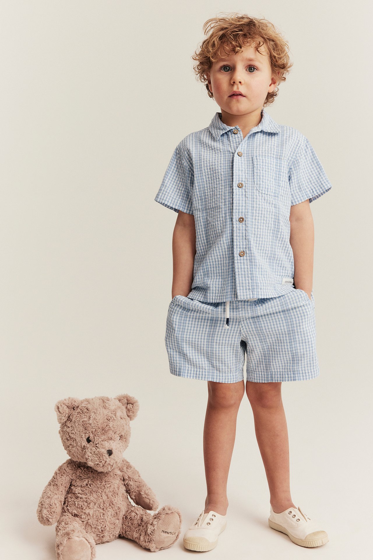 Front view of a kid wearing offwhite checked cotton shorts with a drawstring, a matching shirt, and white shoes.