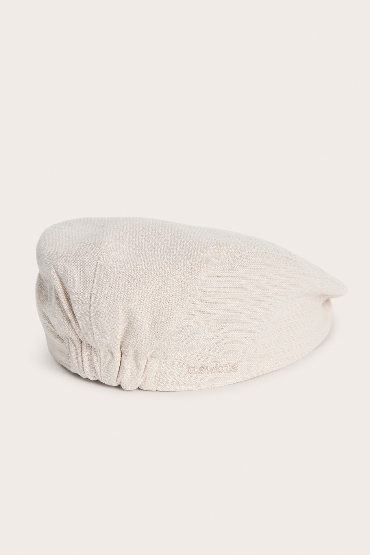 Side view of a beige textured baby/kid's flat cap with elastic back and Newbie embroidery.