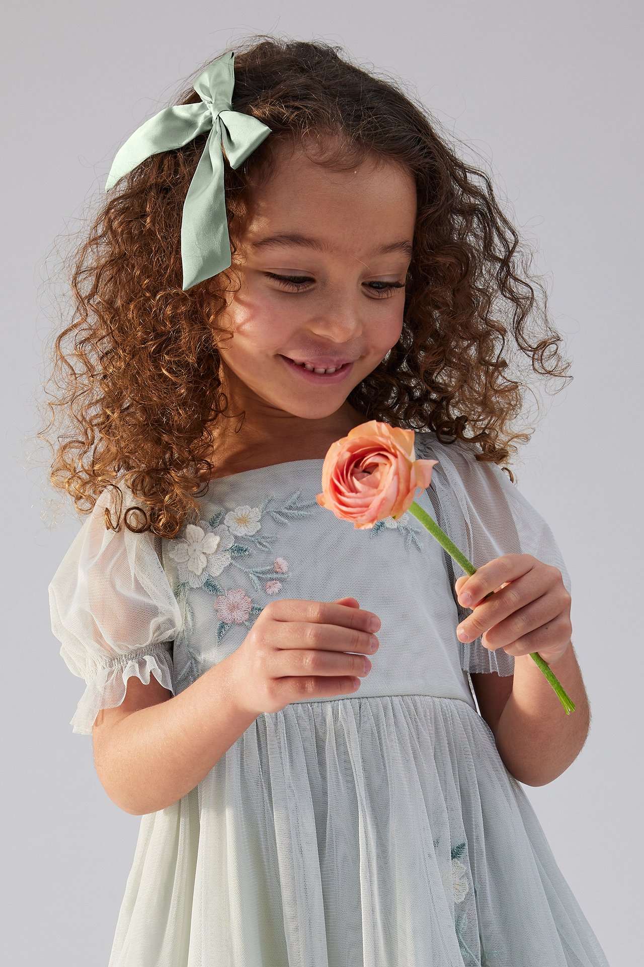 Close-up of a kid wearing a light green satin bow hair clip and a light blue embroidered dress, holding a pink flower.