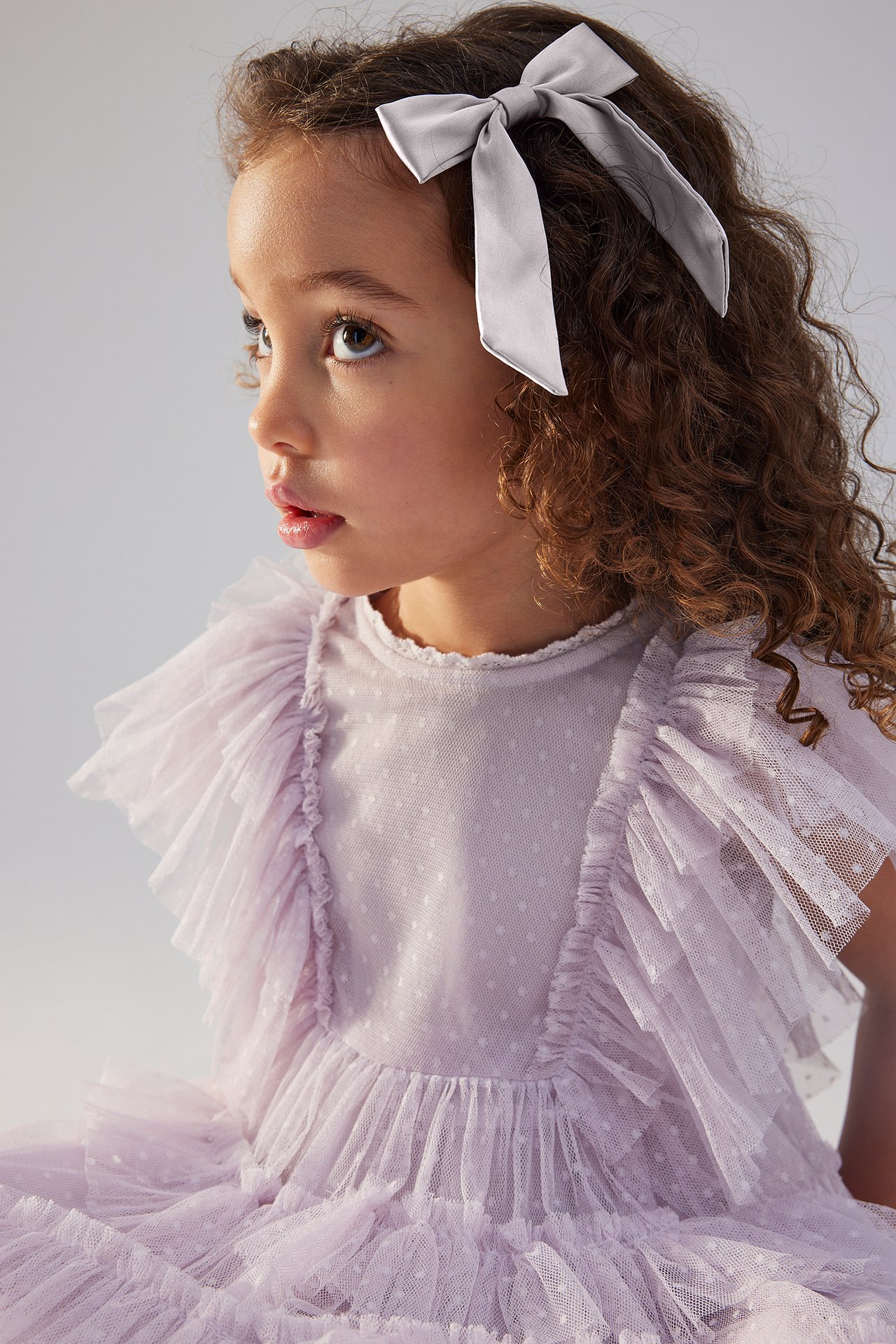Close-up side view of a kid wearing a light lilac satin bow hair clip and a ruffled dotted dress.