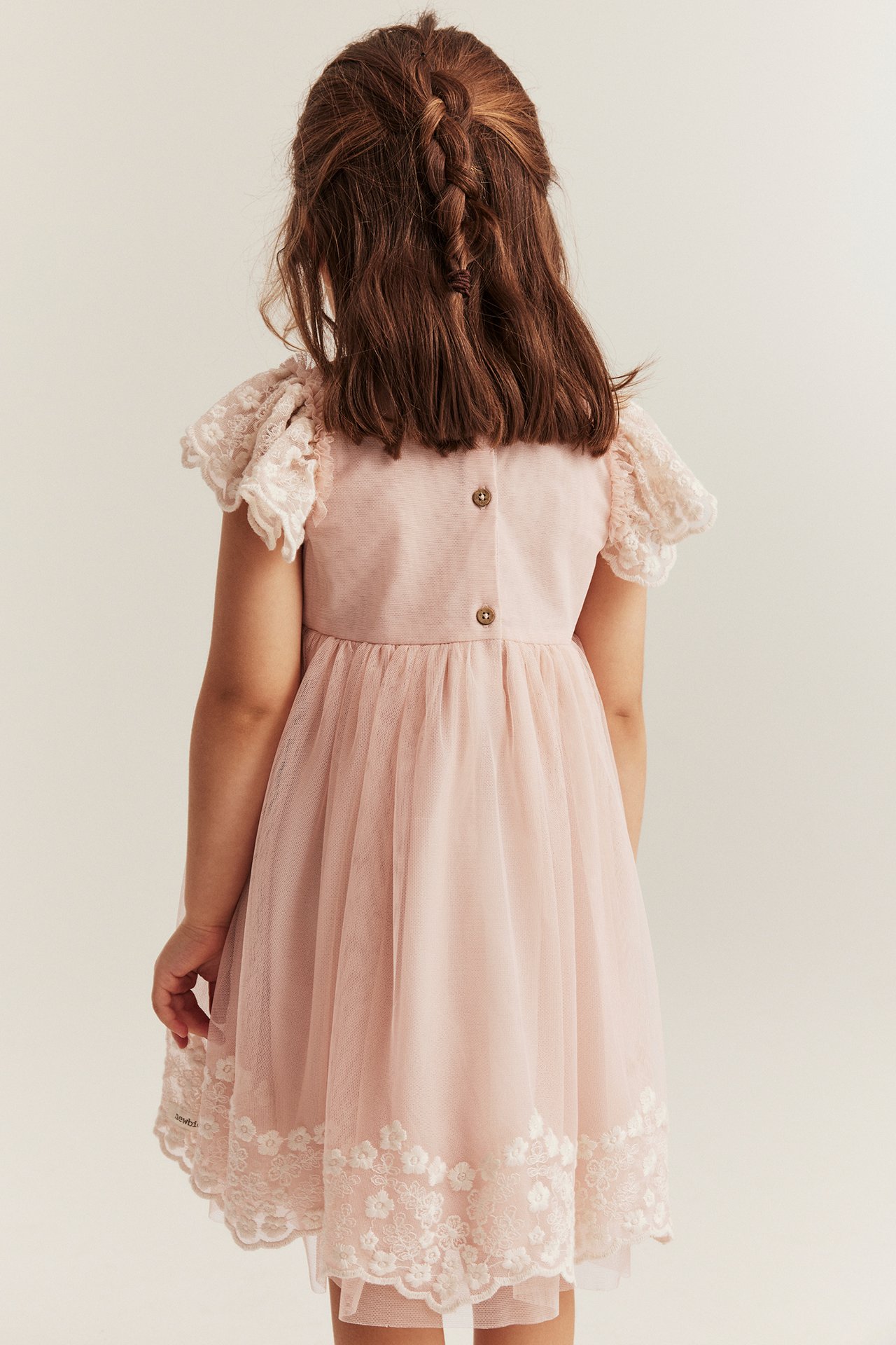 Rückenansicht eines Kindes in einem hellrosa Netz-Kleid mit weißer Blumenstickerei und Knöpfen hinten.
