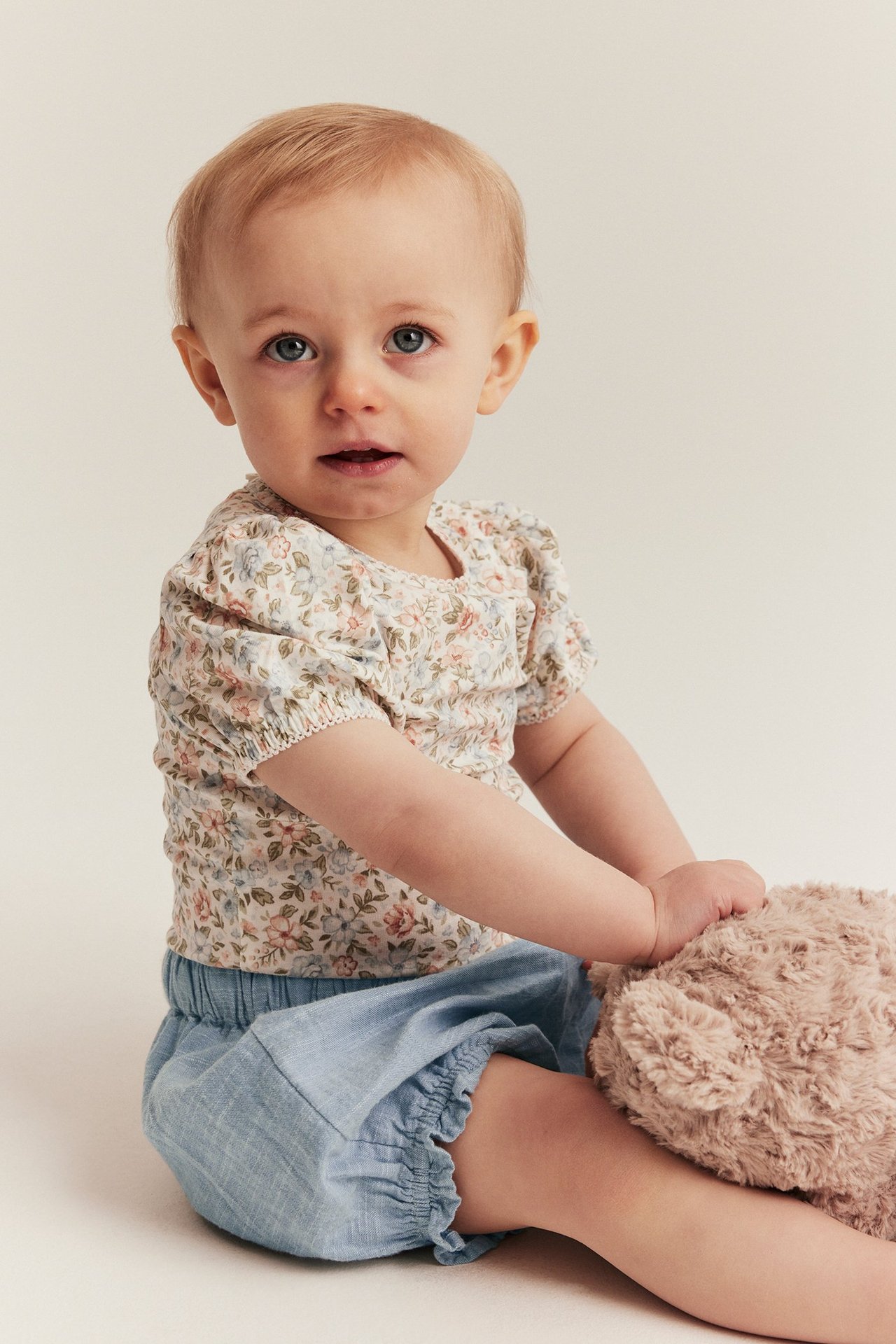 Vorderansicht Baby in hellblauen Rüschen-Chambray-Shorts und floralem Oberteil, hält Teddy.