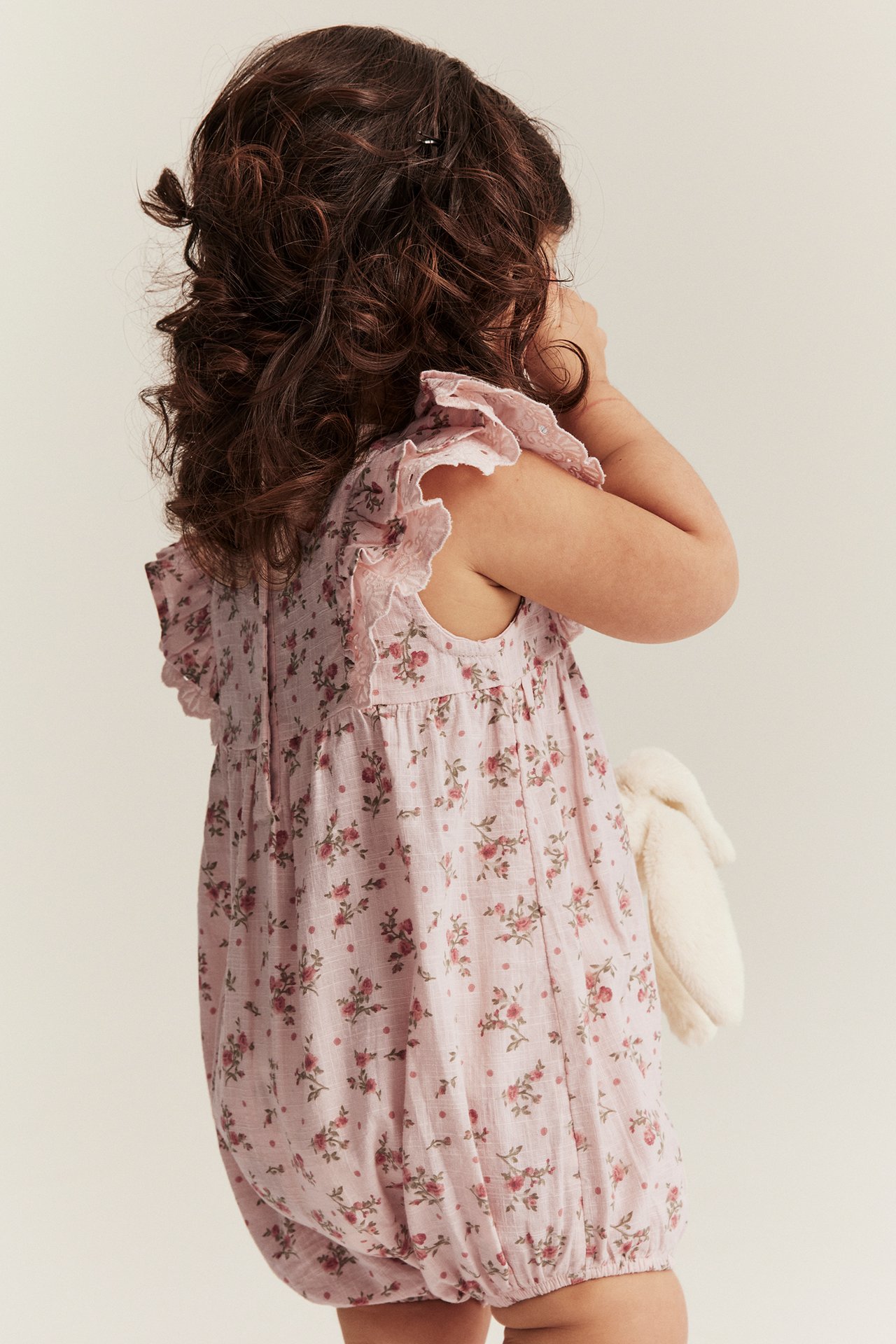 Side view of a baby wearing a light pink floral cotton playsuit with ruffle sleeves, holding a white fluffy toy.